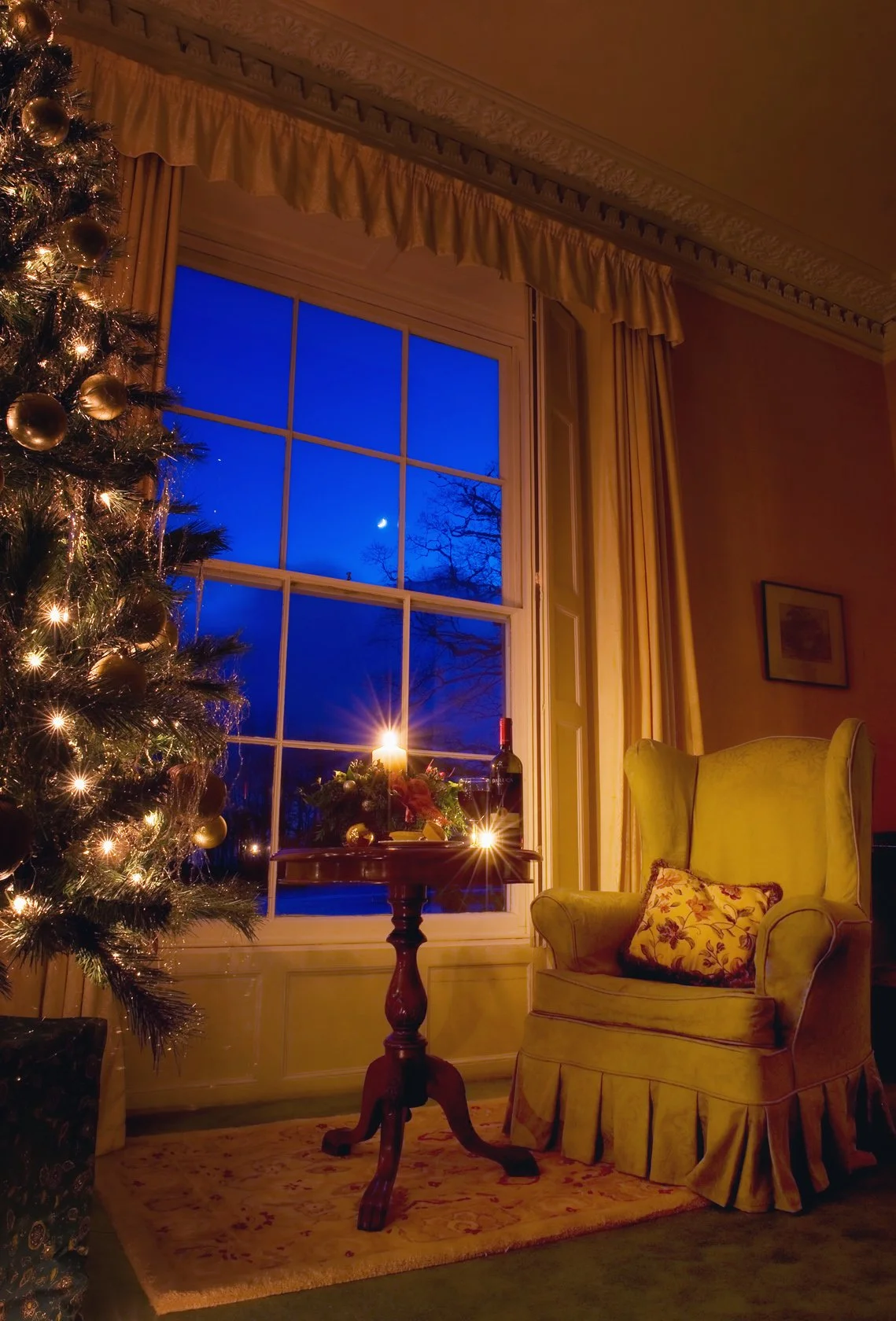 A cozy living room decorated for Christmas with a large decorated Christmas tree, a window with a view of the night sky and a bright crescent moon, a small table with a lit candle and holiday decorations, a green armchair with a floral pillow, and curtains framing the window.