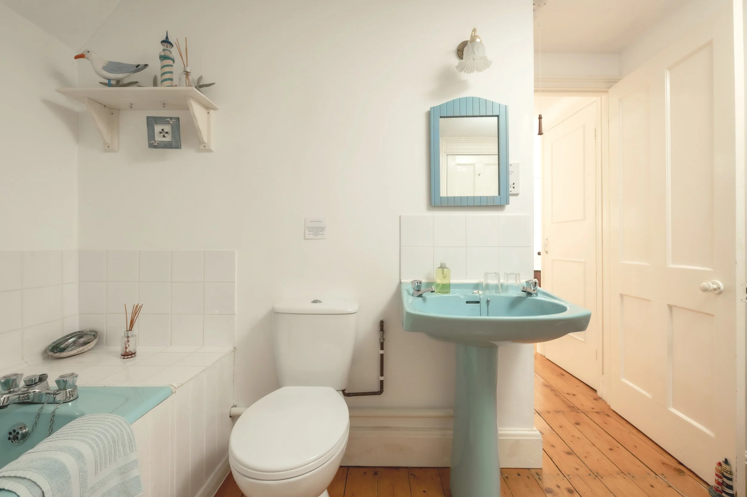 Small vintage bathroom with a light blue sink and mirror, white toilet, wooden floor, and white walls with a shelf holding decorative seagull figure and nautical items at Parnacott house. 
