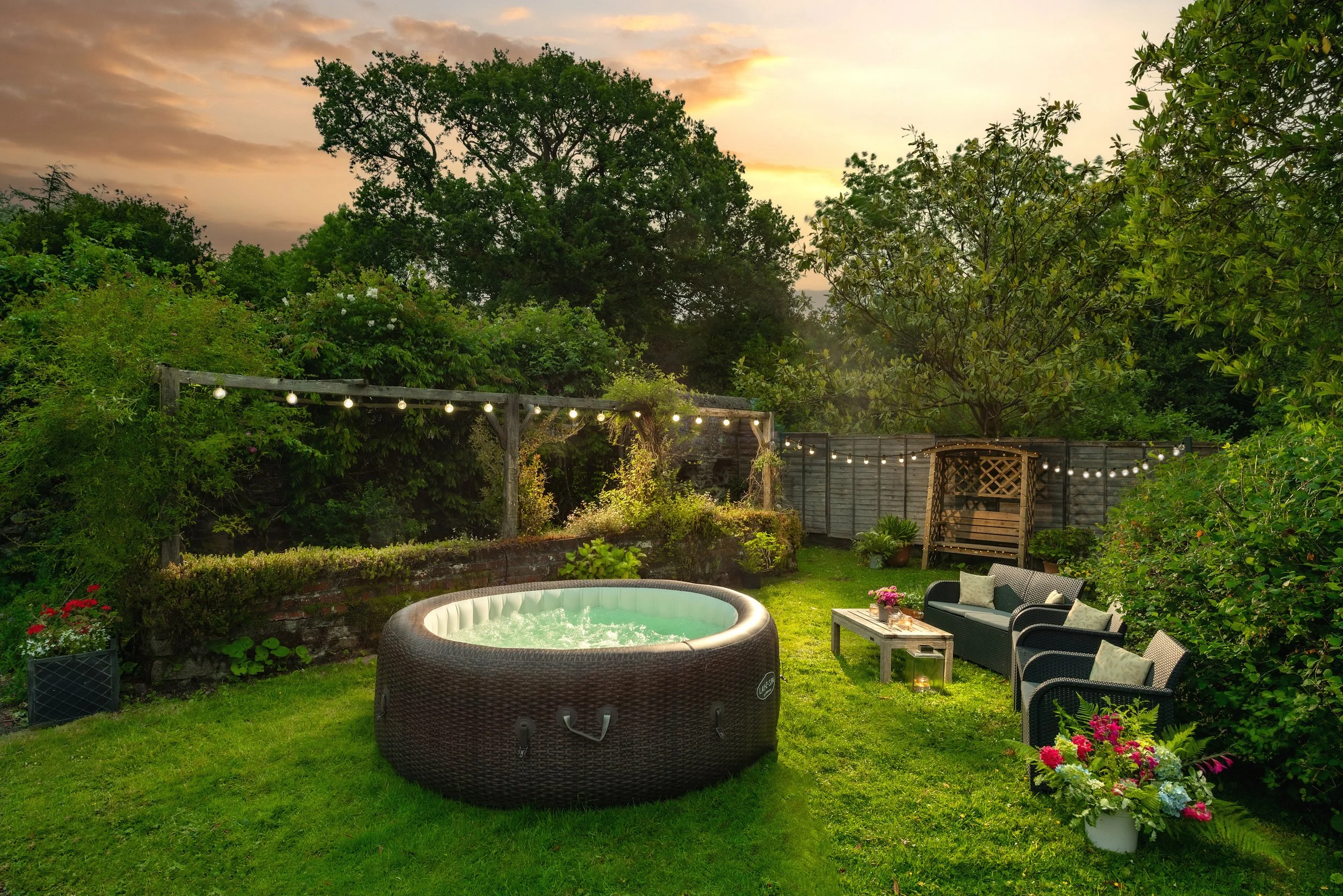 A backyard with a hot tub, outdoor furniture, potted plants, string lights, trees, and a sunset sky at Parnacott house.