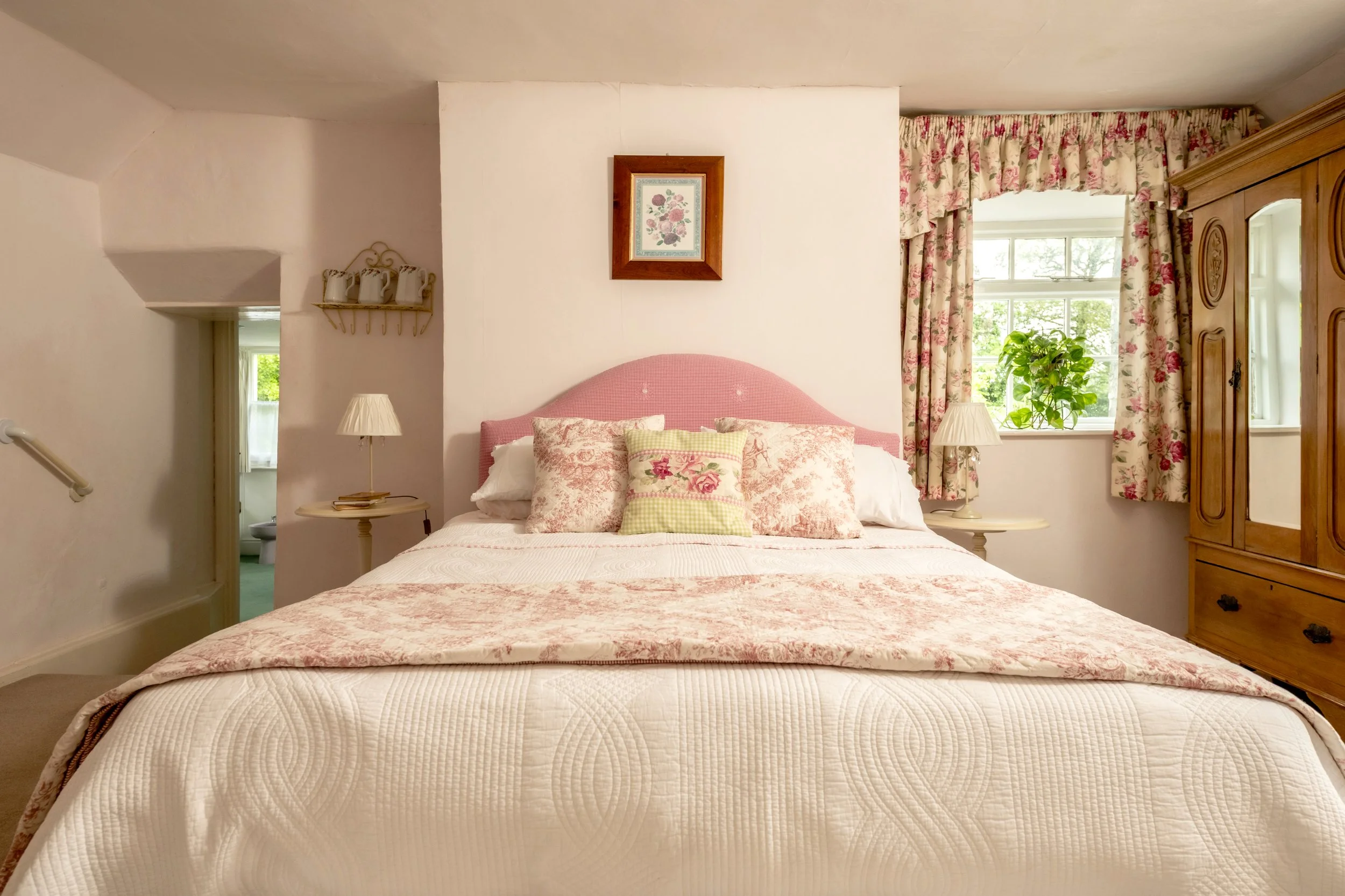 A cozy bedroom with a bed that has pink and floral bedding, a pink headboard, and multiple pillows. There are nightstands with lamps on each side of the bed, floral curtains on the windows, and a wooden wardrobe. A framed floral artwork hangs on the 
