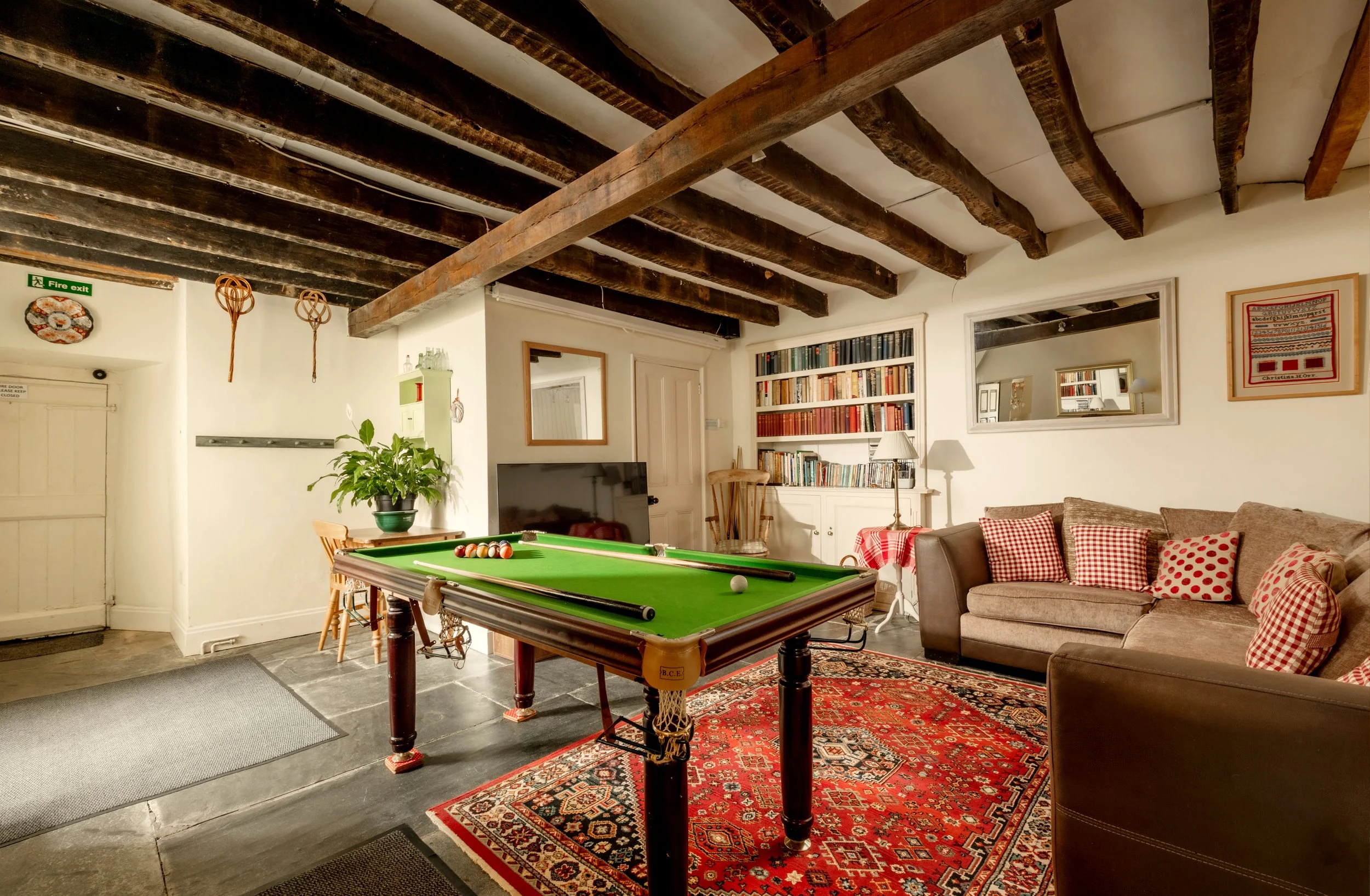 A cozy living room with a green billiards table in the center, surrounded by a patterned red rug and lounging furniture with red and white checkered pillows. There is a bookcase filled with books, a mirror, a lamp, and decorative items on the wall, with wooden ceiling beams overhead.