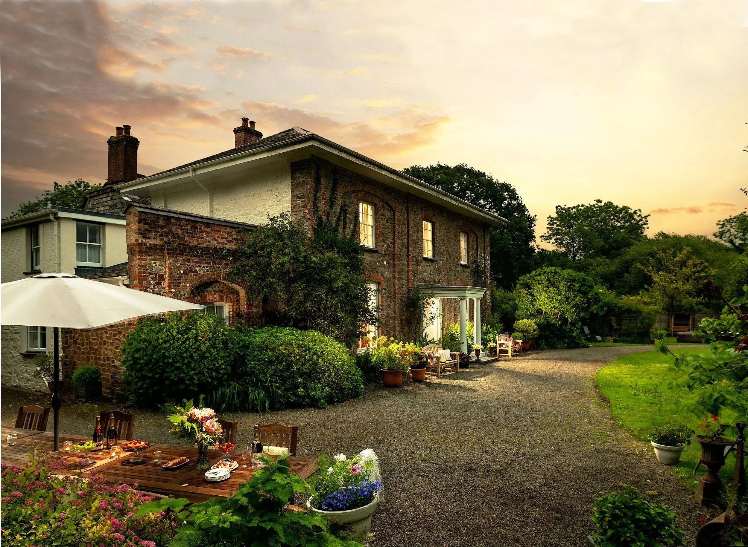 A cozy outdoor garden with a brunch setup featuring a variety of dishes and drinks on a wooden table under a large umbrella. The garden has lush greenery & potted plants, with charming brick house in the background during sunset at Parnacott House.