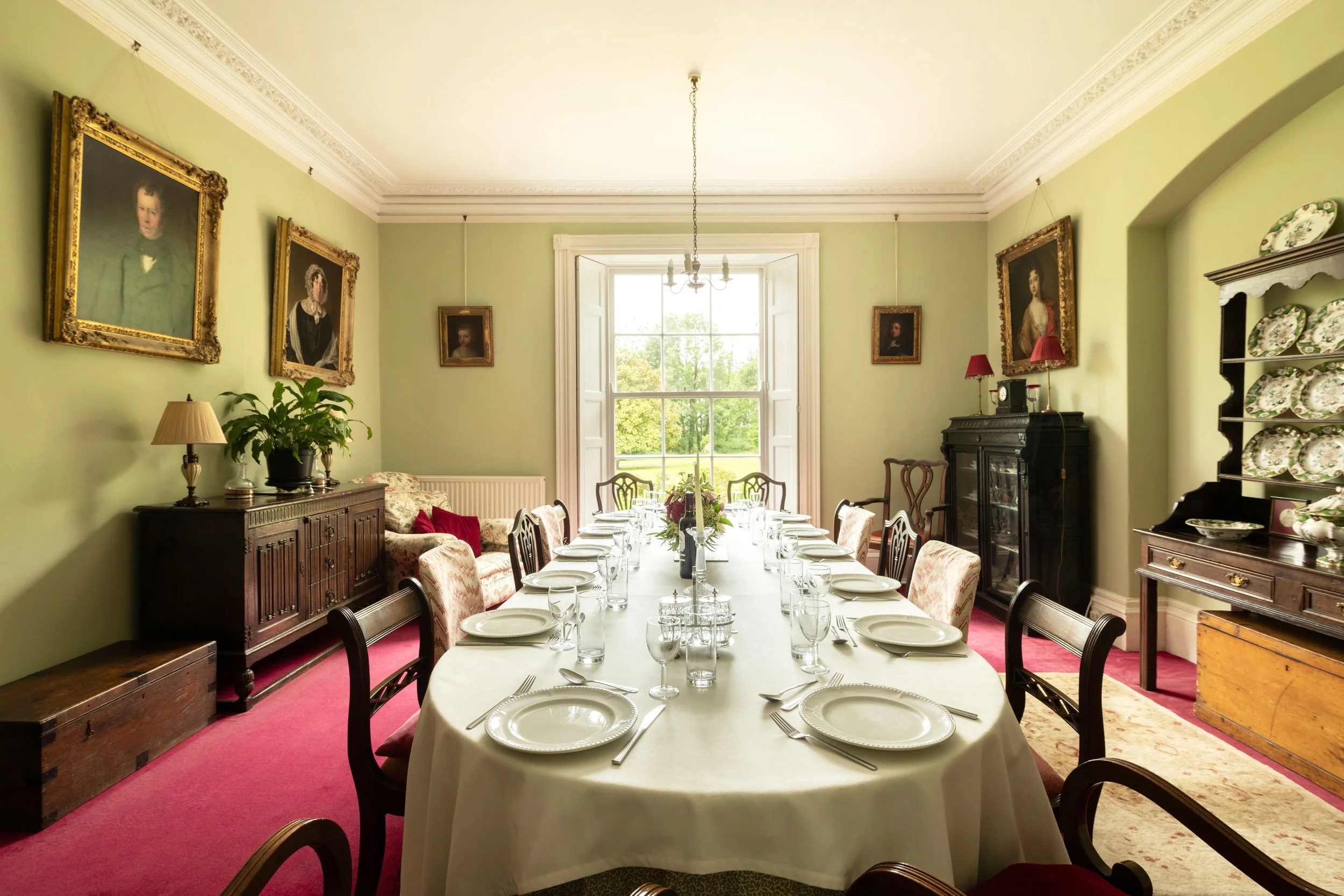 A dining room with a long table set with plates, glasses, and cutlery, surrounded by chairs. The walls are light green with framed portraits and a large window with white shutters in the background. There are antique furniture pieces, including a dark wooden sideboard, a display cabinet with decorative plates, and a fireplace mantel. The ceiling has decorative molding and a simple chandelier.