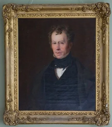 A framed classical portrait painting of a man with short, curly brown hair, wearing a black coat, white shirt, and black cravat, against a dark background at Parnacott house.
