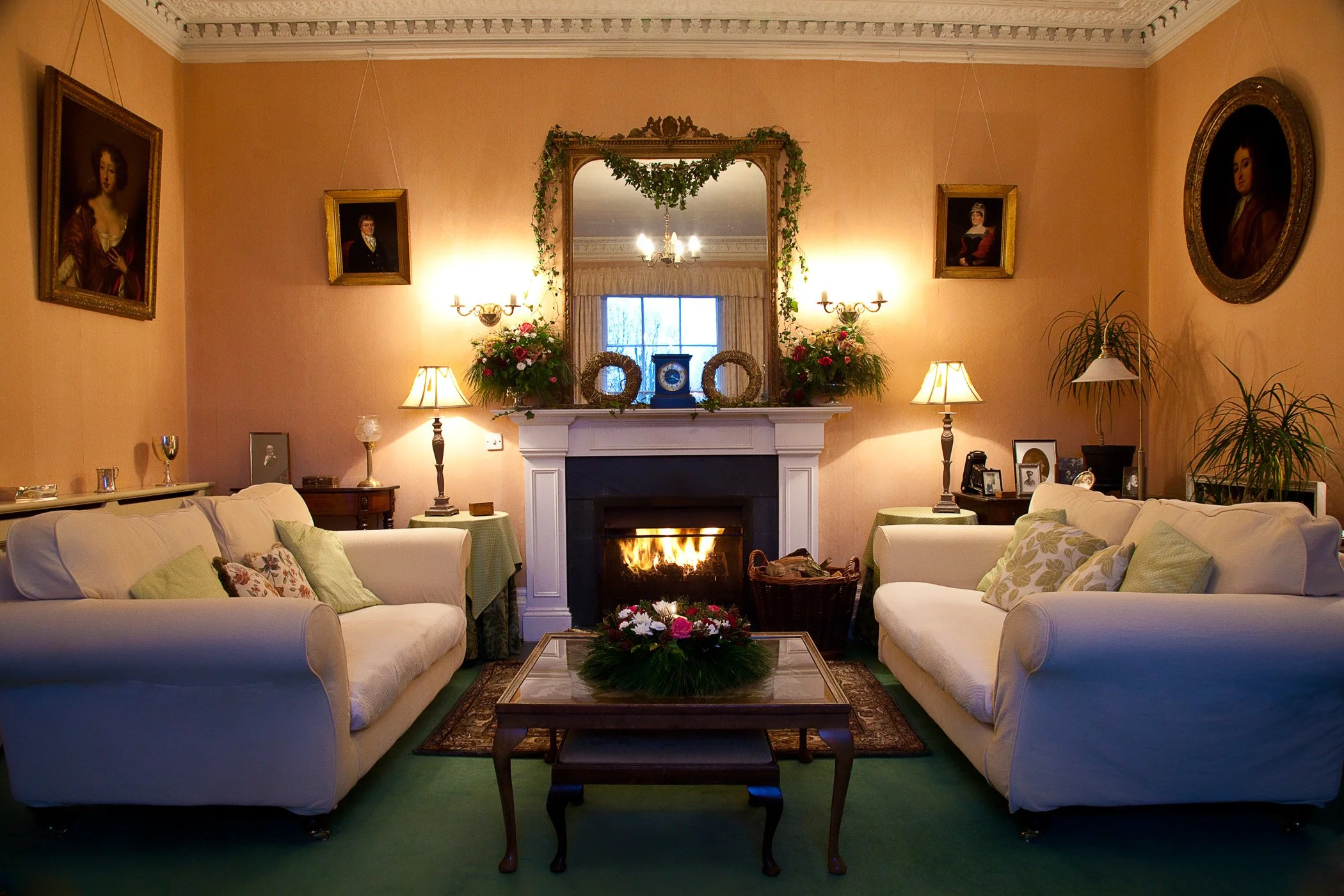 A cozy living room with two white sofas, a coffee table with a flower arrangement, a fireplace with a mirror above, candle sconces, and framed portraits on the peach-colored walls. There are also various lamps and plants at Parnacott house.