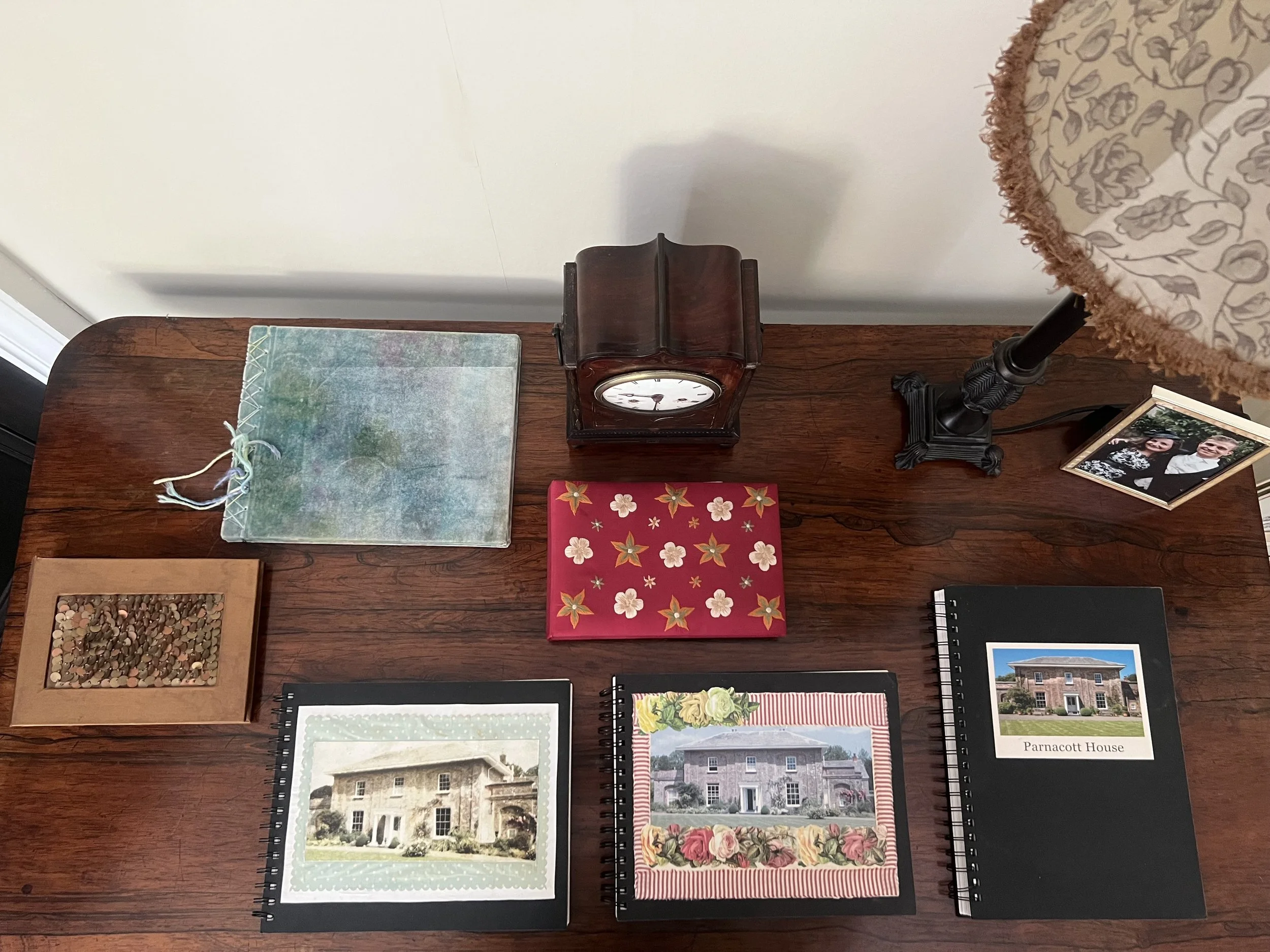 A wooden desk with several books, photos, and decorative items, including an analog clock with a wooden frame, a table lamp with a floral-patterned shade, and framed photographs, one labeled Parncutt House.