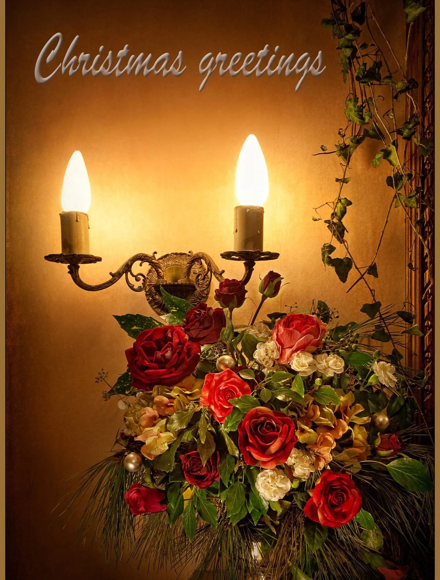 A floral arrangement with red, cream, and peach roses, green leaves, and decorative gold ornaments, beneath a vintage gold wall sconce with two lit candles, against a beige background. The words 'Christmas greetings' are written in gray script at the top left of the image.