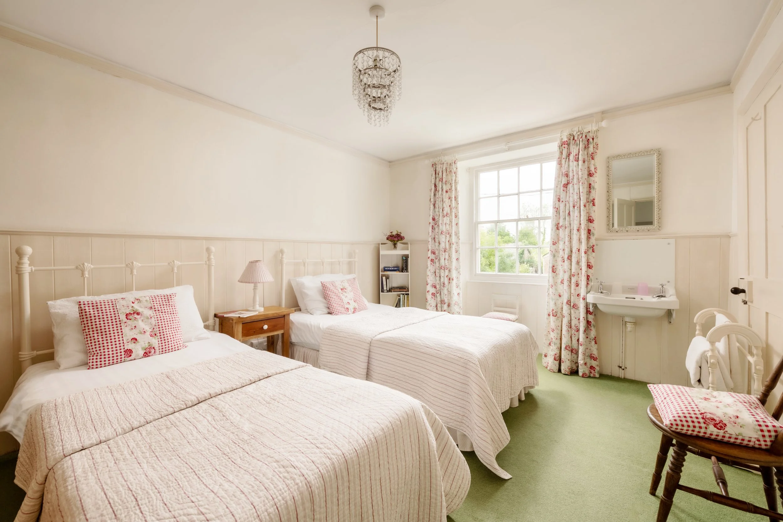A bedroom with four white beds, each with pink and white checkered pillows, a bedside table with a lamp, a window with floral curtains, and a mirror on the wall. There is a small sink and a rocking chair with a matching checkered cushion. The room ha
