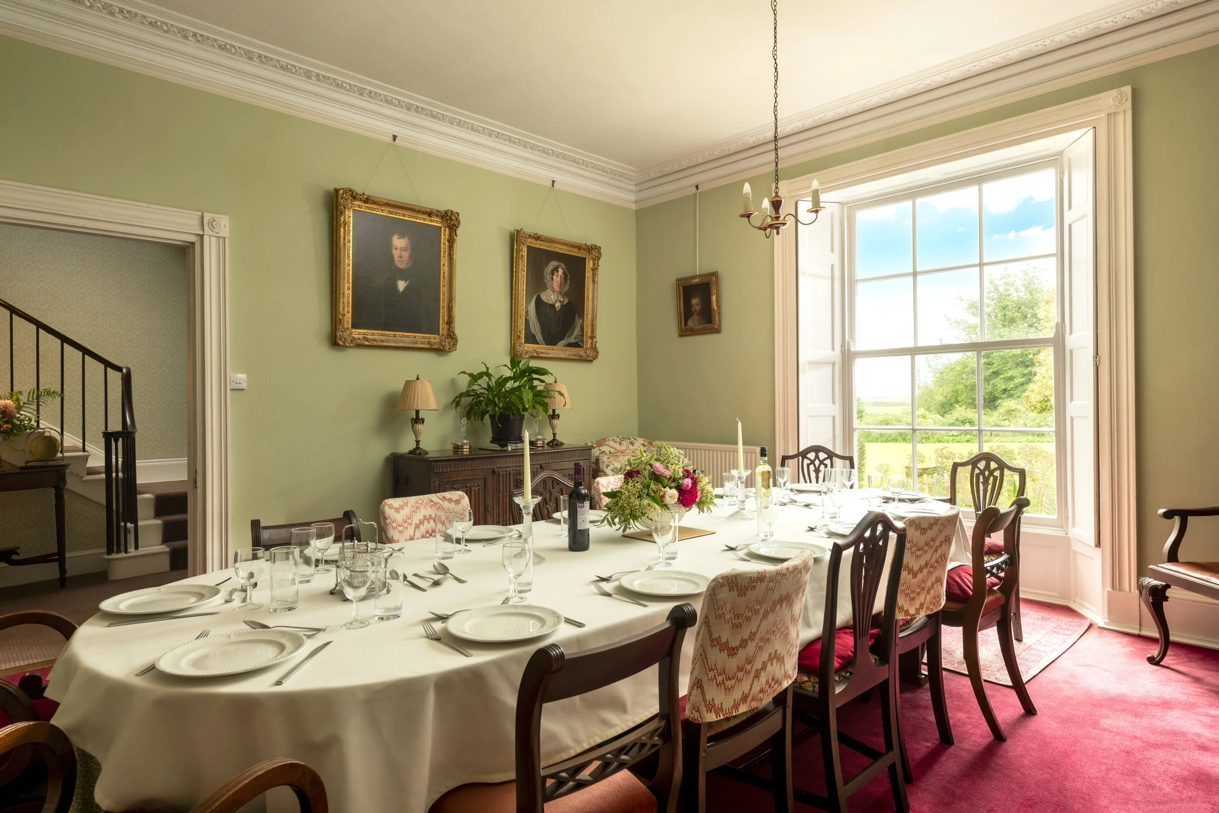 Dining room with a large window, decorated with portraits, a chandelier, and a table set with glassware and floral centerpiece at Parnacott house.