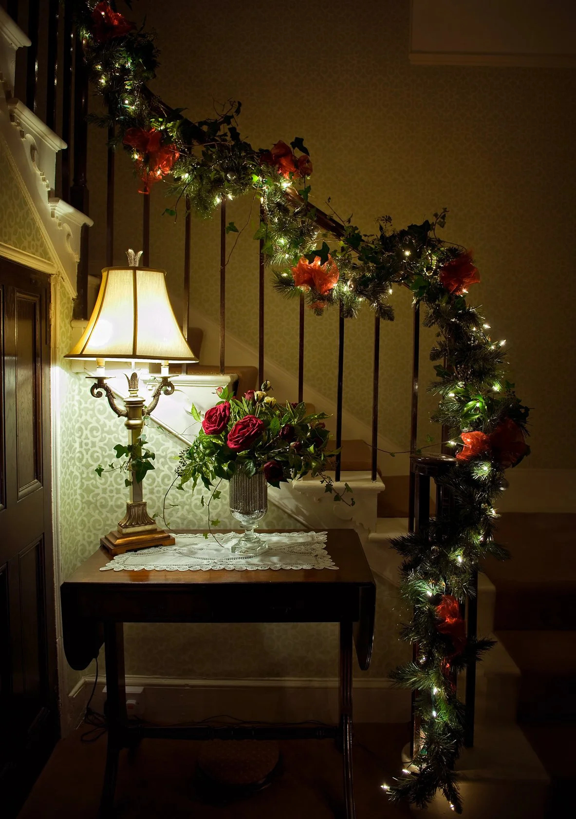 Decorated staircase with greenery, red flowers, and string lights, along with a table holding a vase of red roses and a lit table lamp at Parnacott house.