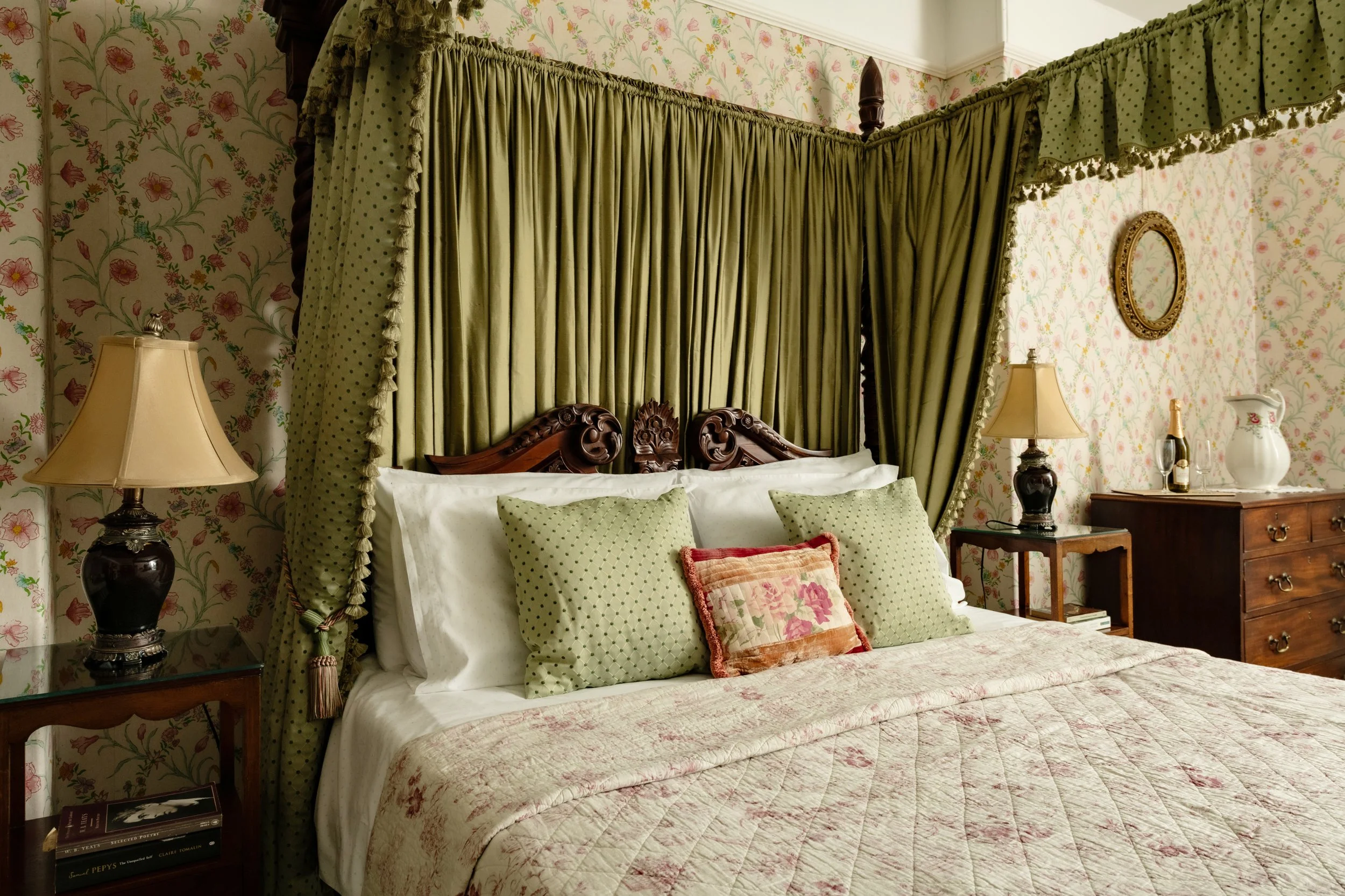 A bedroom with floral wallpaper, green curtains surrounding a four-poster bed with wooden carving details and green polka dot pillows, a nightstand with a lamp, and decorative items including a mirror and bottles at Parnacott house. 