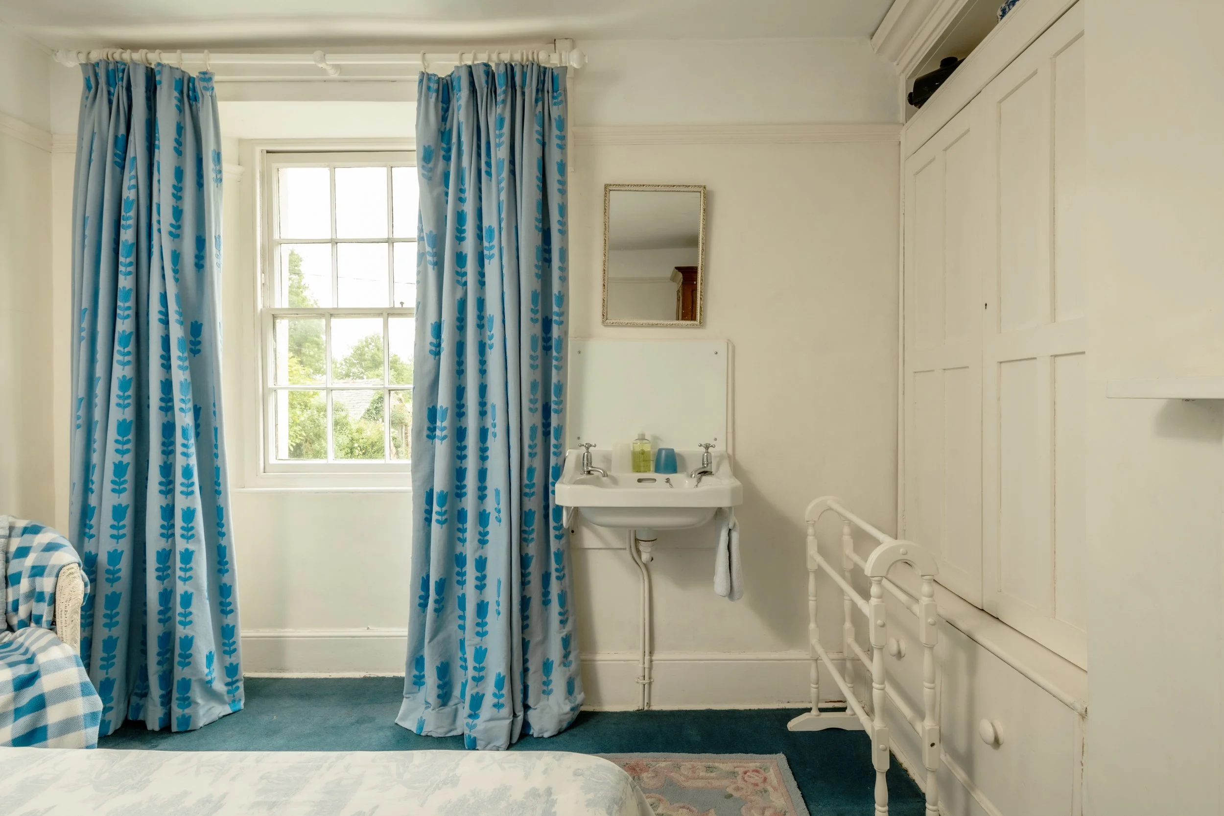 A bedroom with a window and white walls, decorated with blue curtains featuring a leaf pattern, a small white sink with a mirror above it, and a white bed frame on the right side of the image at Parnacott house. 