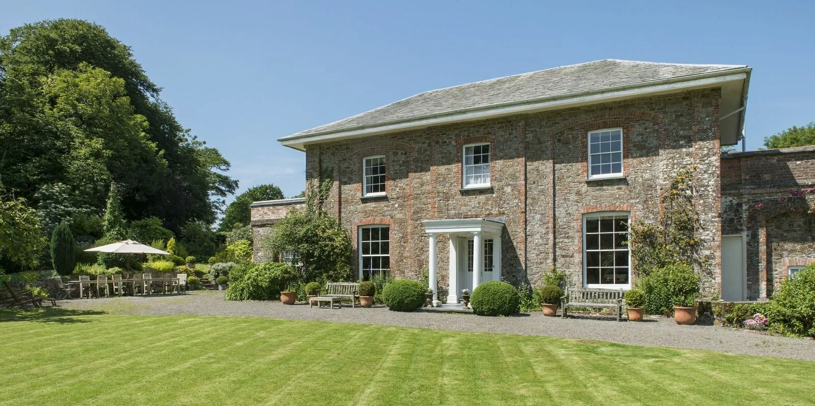 A large brick house with multiple windows and a small porch, surrounded by a well-maintained garden with benches, potted plants, and a grassy lawn under a clear blue sky at Parnacott house.