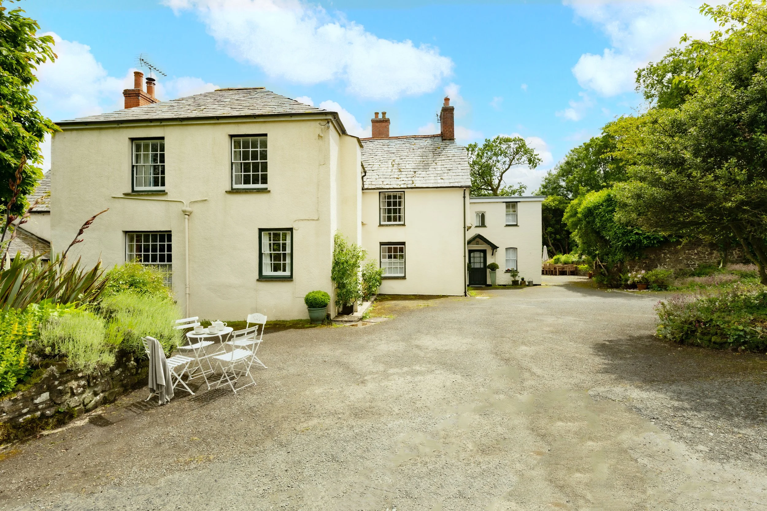 A charming white house with multiple windows and a dark front door, surrounded by lush greenery and a gravel courtyard. There's outdoor furniture with a small table and chairs on the left side at Parnacott house.