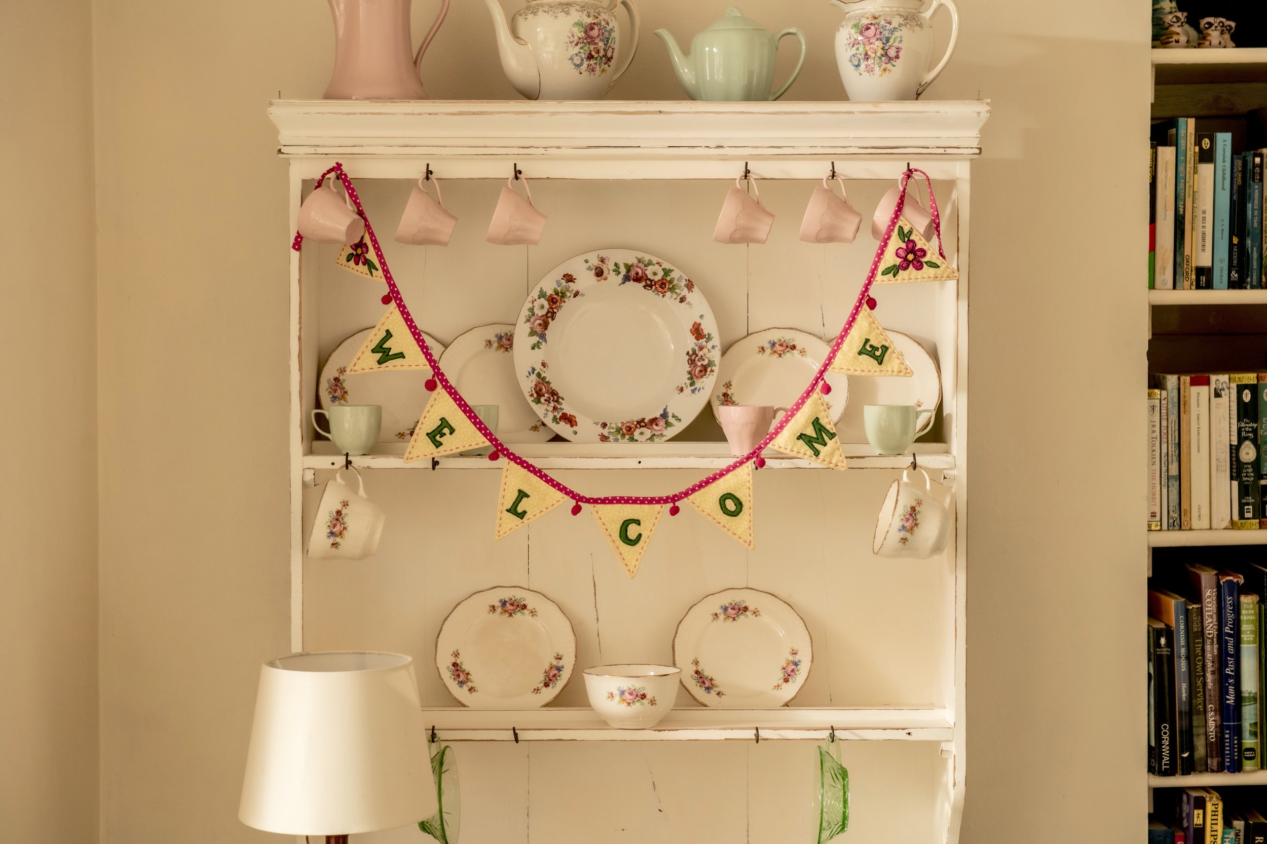 Decorative white bookcase with vintage floral china plates, cups, and teapots, along with hanging teacups and a colorful banner spelling 'WELCOME'; books on the side at Parnacott house.