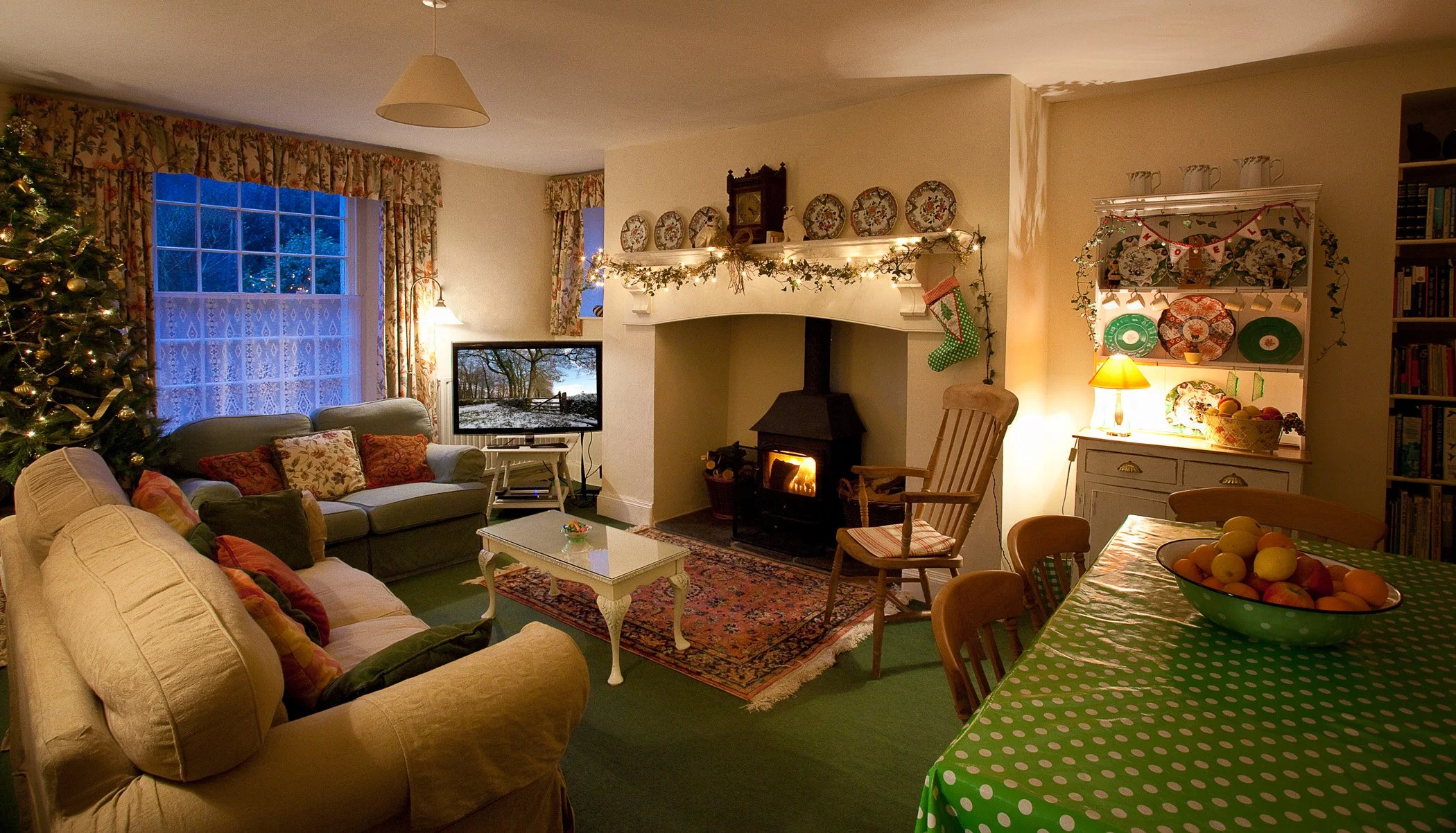 A cozy living room decorated for Christmas with a Christmas tree, stockings, and garlands. There is a fireplace with a fire, a sofa with pillows, a table, and a dining area with a green polka-dot tablecloth and a fruit bowl at Parnacott house.