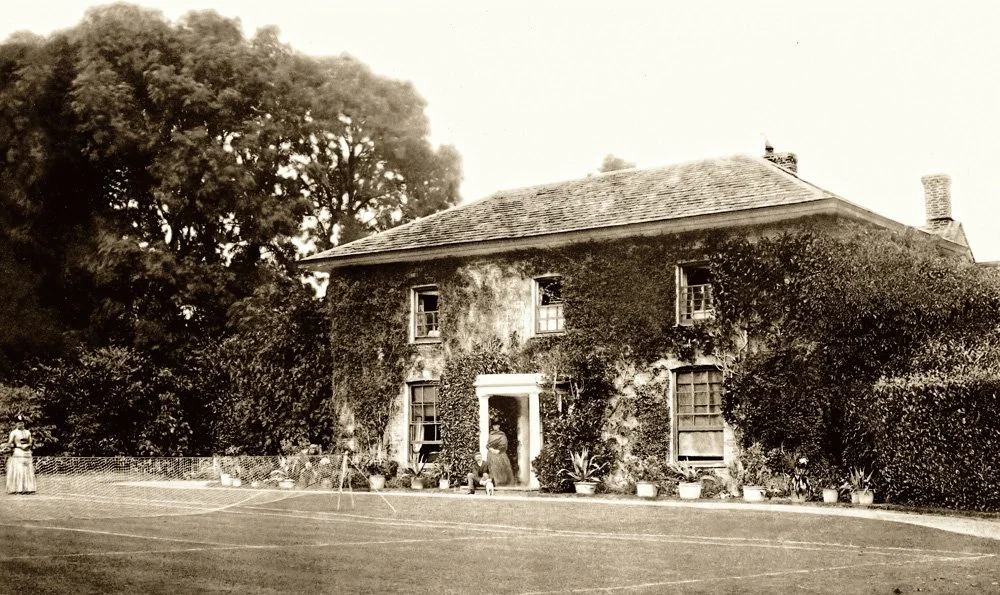 A woman standing on a tennis court near an old ivy-covered house in the daytime 1880's at Parnacott house.