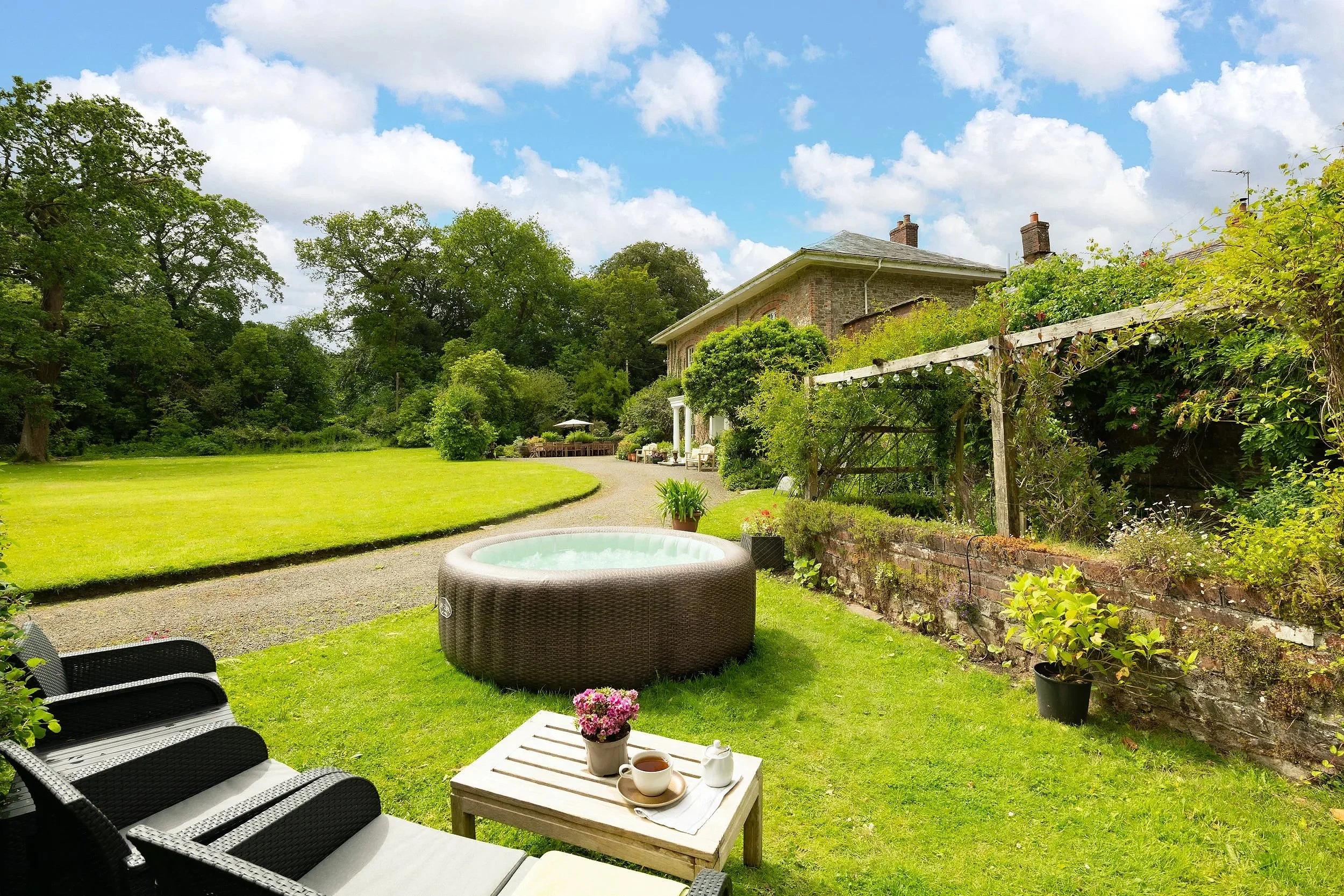 A backyard garden with a lawn, patio, outdoor seating, potted plants, a hot tub, and a brick house with a garden behind it at Parnacott house.