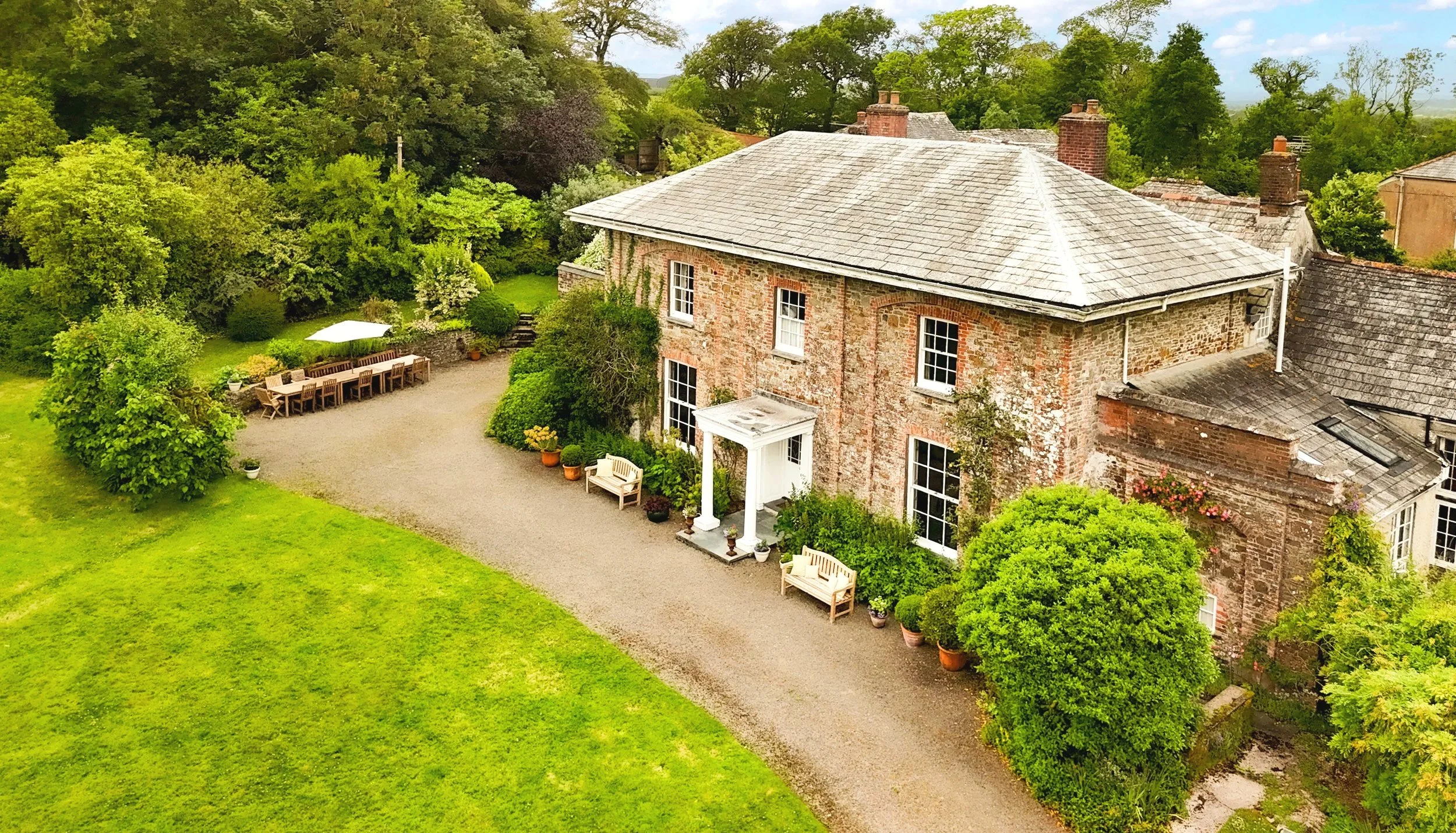Aerial view of a brick house with a grey roof surrounded by lush green trees and grass, with an outdoor dining area and benches in the garden at Parnacott house.