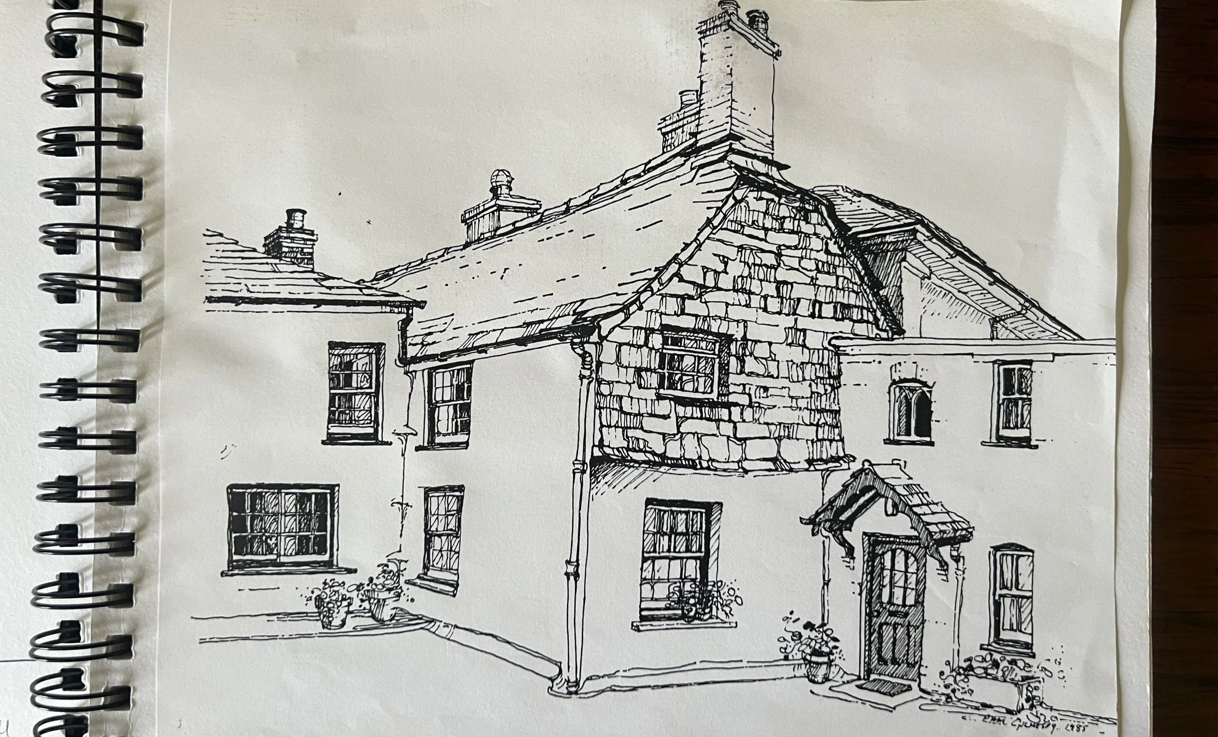 A black and white ink drawing of a traditional brick house with multiple windows, a sloped roof, chimneys, and a small porch, with potted plants and flowers outside at Parnacott house.