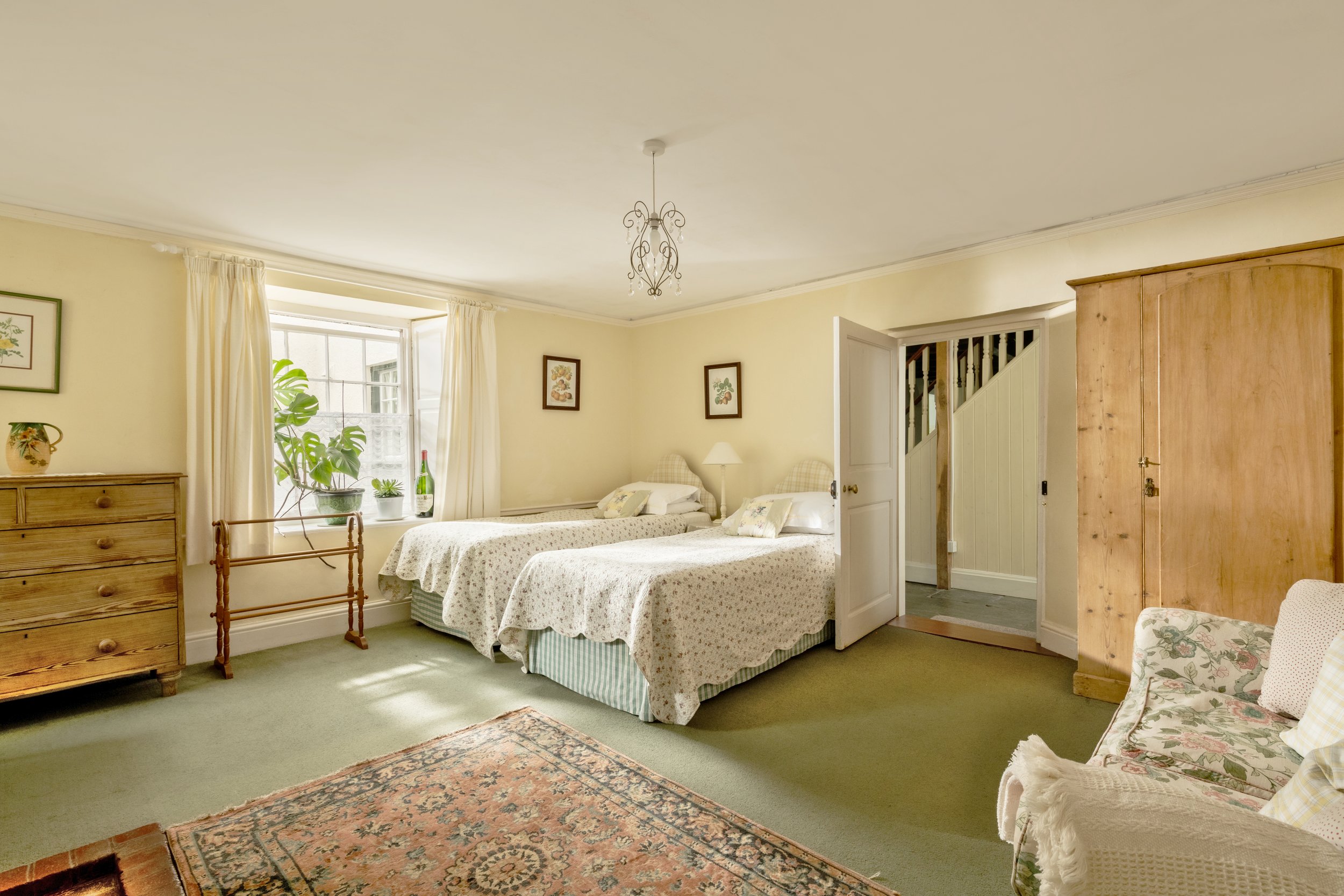 A cozy bedroom with two twin beds with floral bedspreads, a wooden dresser, a window with cream curtains, potted plants on the windowsill, framed artwork on the walls, a chandelier hanging from the ceiling, and a patterned rug on the carpeted floor.