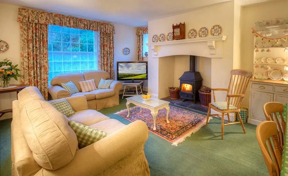 Cozy living room with cream sofas, floral curtains, a fireplace with a wood stove, a TV, and a white coffee table on a colorful rug at Parnacott house.