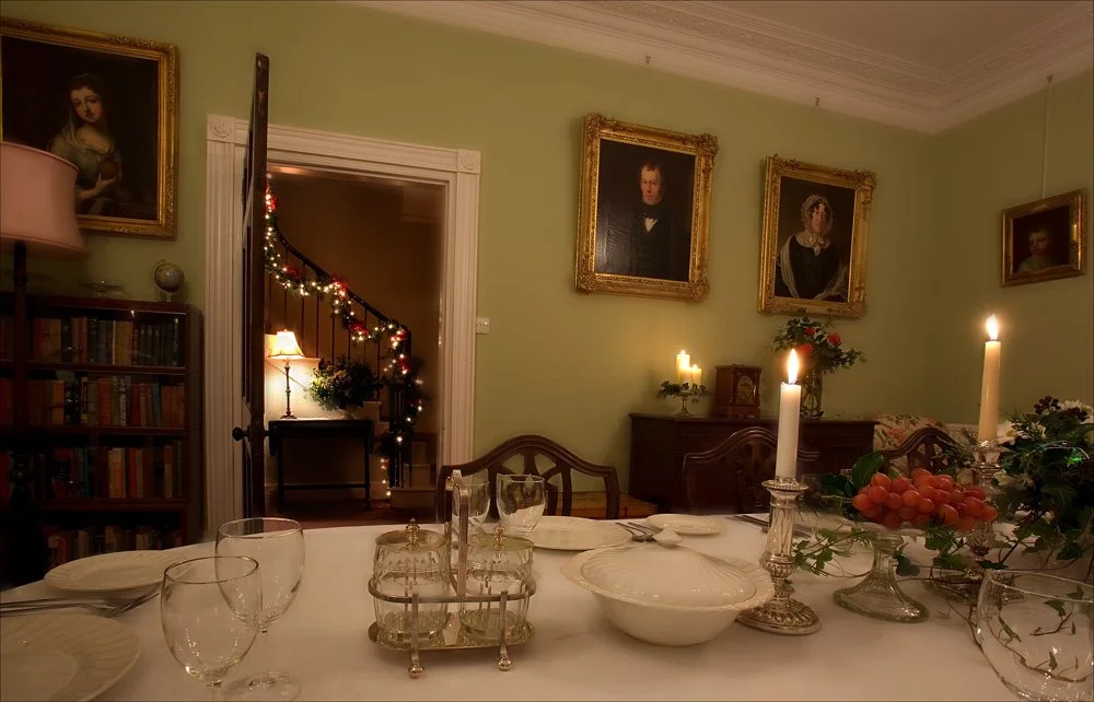 A dining room with a table set for a formal meal, decorated with candles, flowers, and grapes. The room has light green walls and framed portraits hanging on the walls. In the background, there is a door leading to a staircase decorated with Christmas lights and a small table with a lamp.