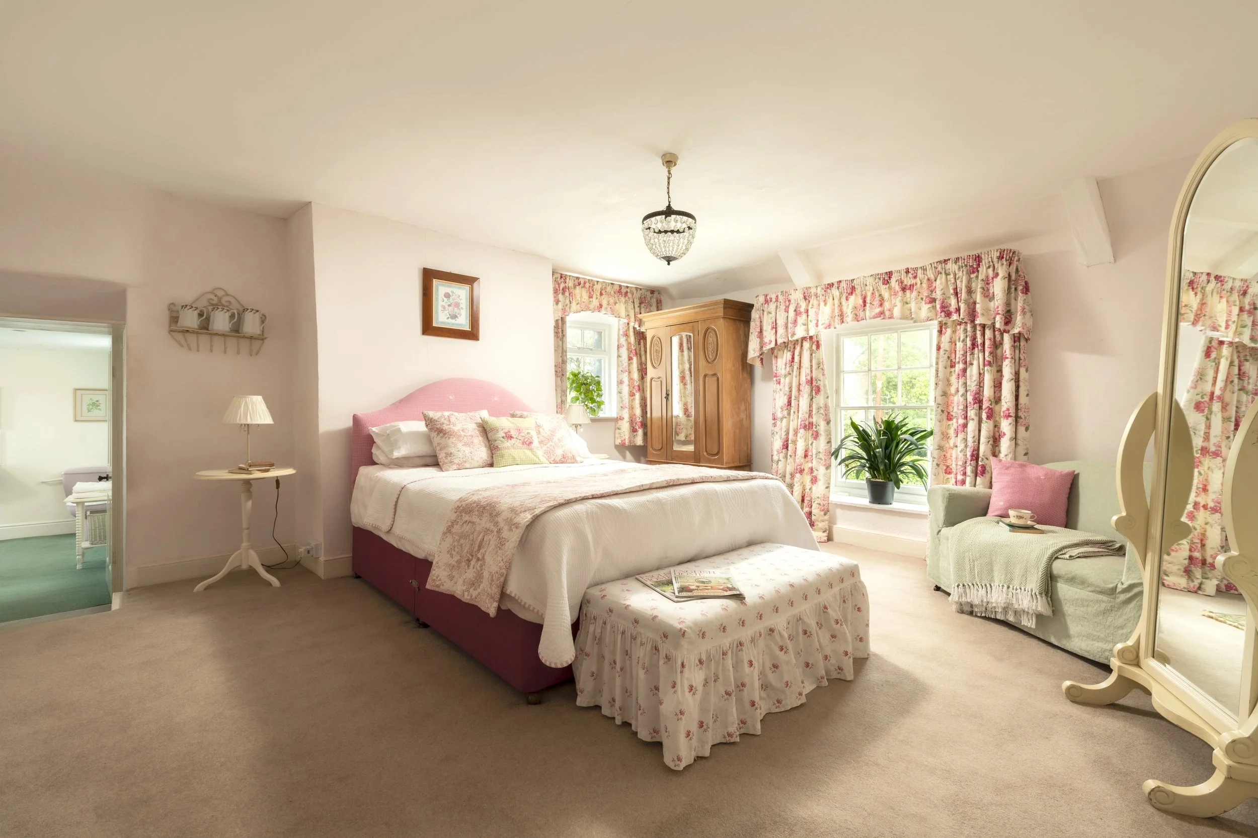 A cozy bedroom with a pink upholstered bed, floral curtains, a wooden wardrobe, a small side table with a lamp, and a curved mirror. There is also a green armchair with pink cushions and a plant near the windows at Parnacott house. 