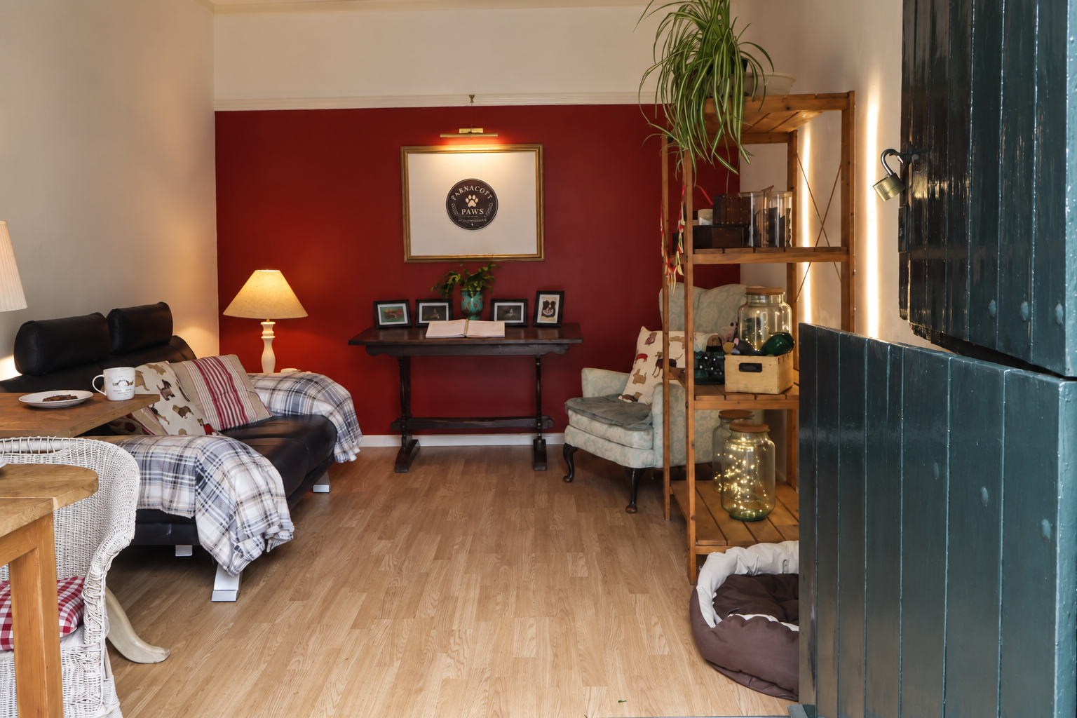 Living room with a black sofa, lamp, a red accent wall with framed pictures, a wooden table, a light-colored armchair with dog-themed pillows, tall wooden shelf with jars and decorative items, & dog bed on the floor. Parnacott house. Parnacott Paws.