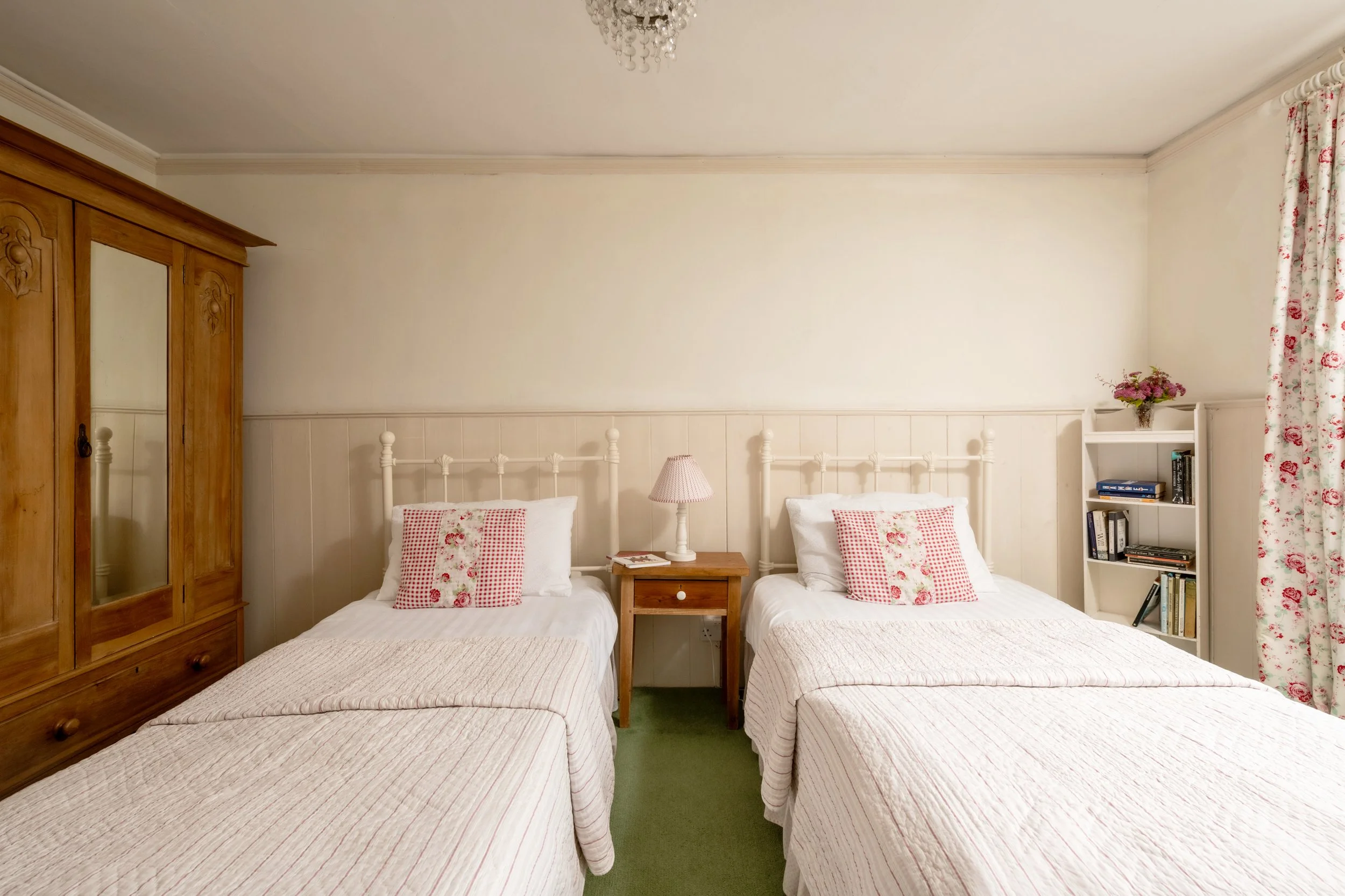 A bedroom with three single beds, each with white bedding and red-patterned pillows. A wooden wardrobe with mirrored doors is on the left, and a small bedside table with a pink lampshade is between two of the beds. On the right, a white bookshelf hol