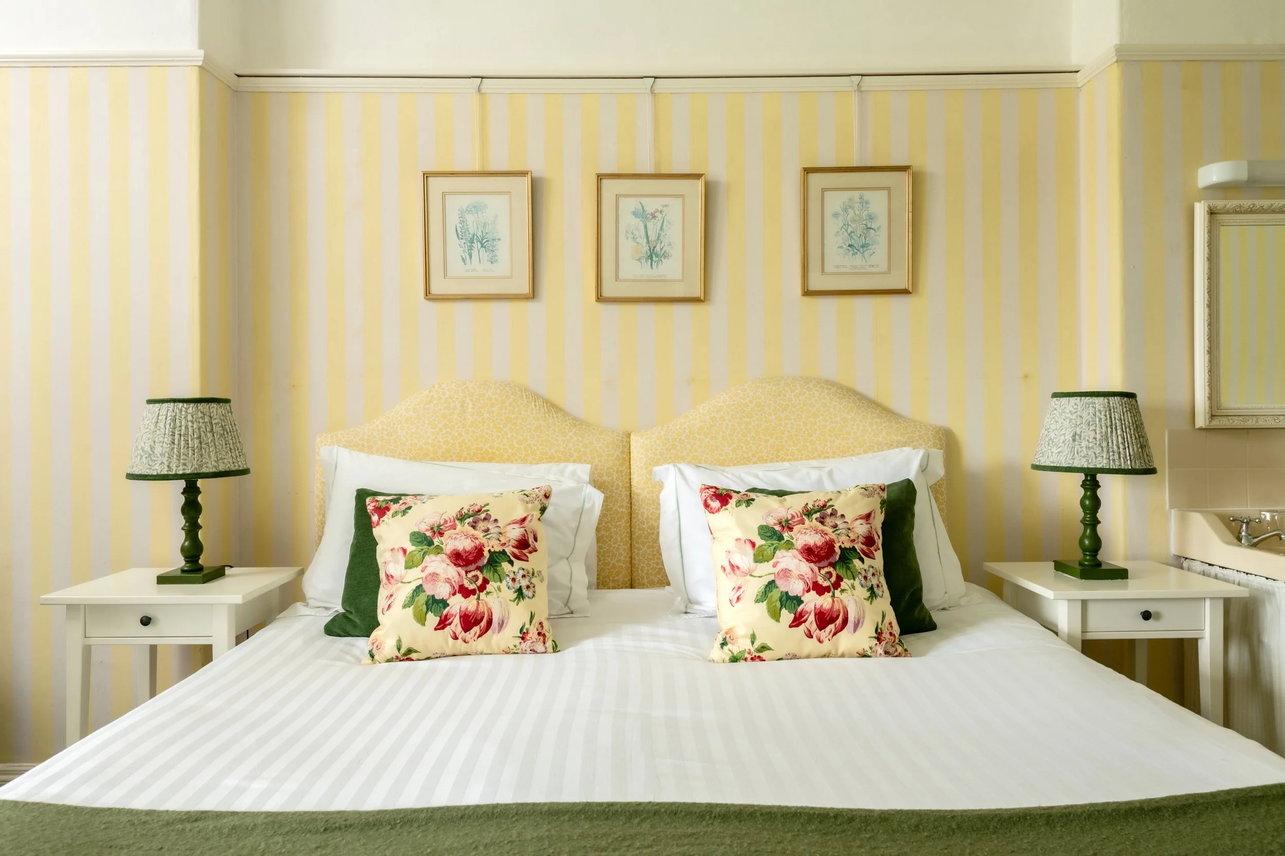 A bedroom with yellow striped wallpaper, two nightstands with green lamps, a bed with floral pillows, and framed botanical prints above the headboard at Parnacott house. 