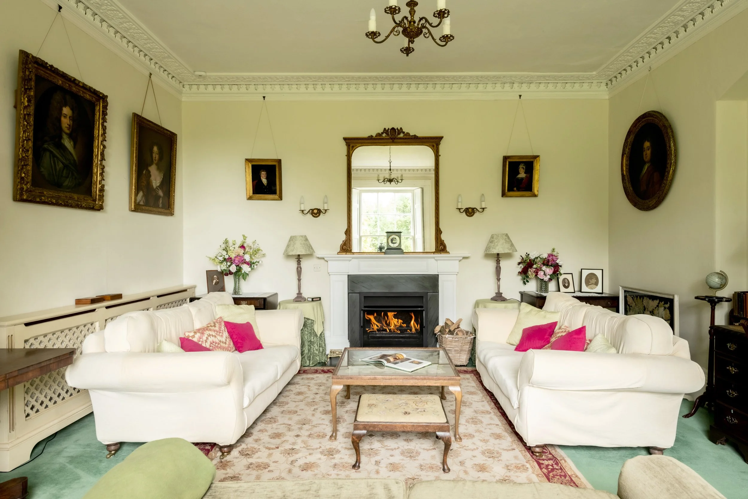 A living room with two white sofas, a fireplace with a mirror above it, and floral arrangements. Five framed portraits are on the cream-colored walls, and there is an ornate chandelier hanging from the ceiling at Parnacott house.