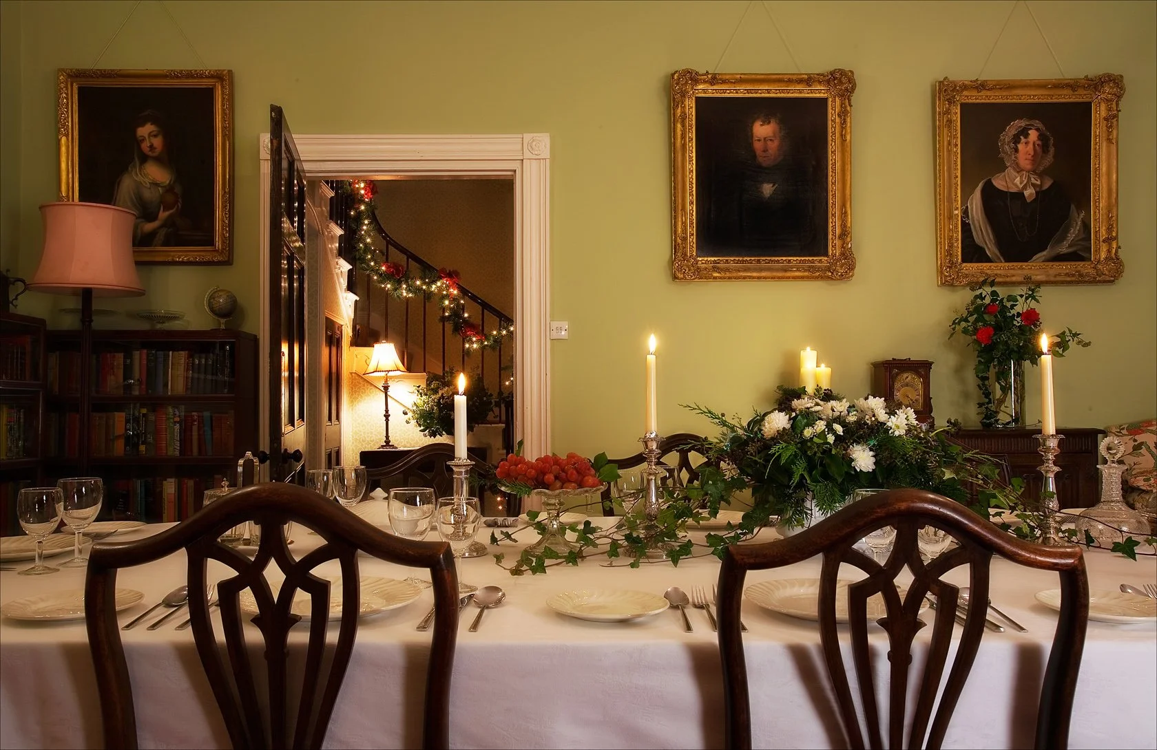 A decorated dining room with a long table set for a meal, featuring candles, flowers, and grapes as centerpieces. The room has a green wall with three framed classical portraits, a bookshelf, and a doorway leading to a staircase decorated with Christmas lights.