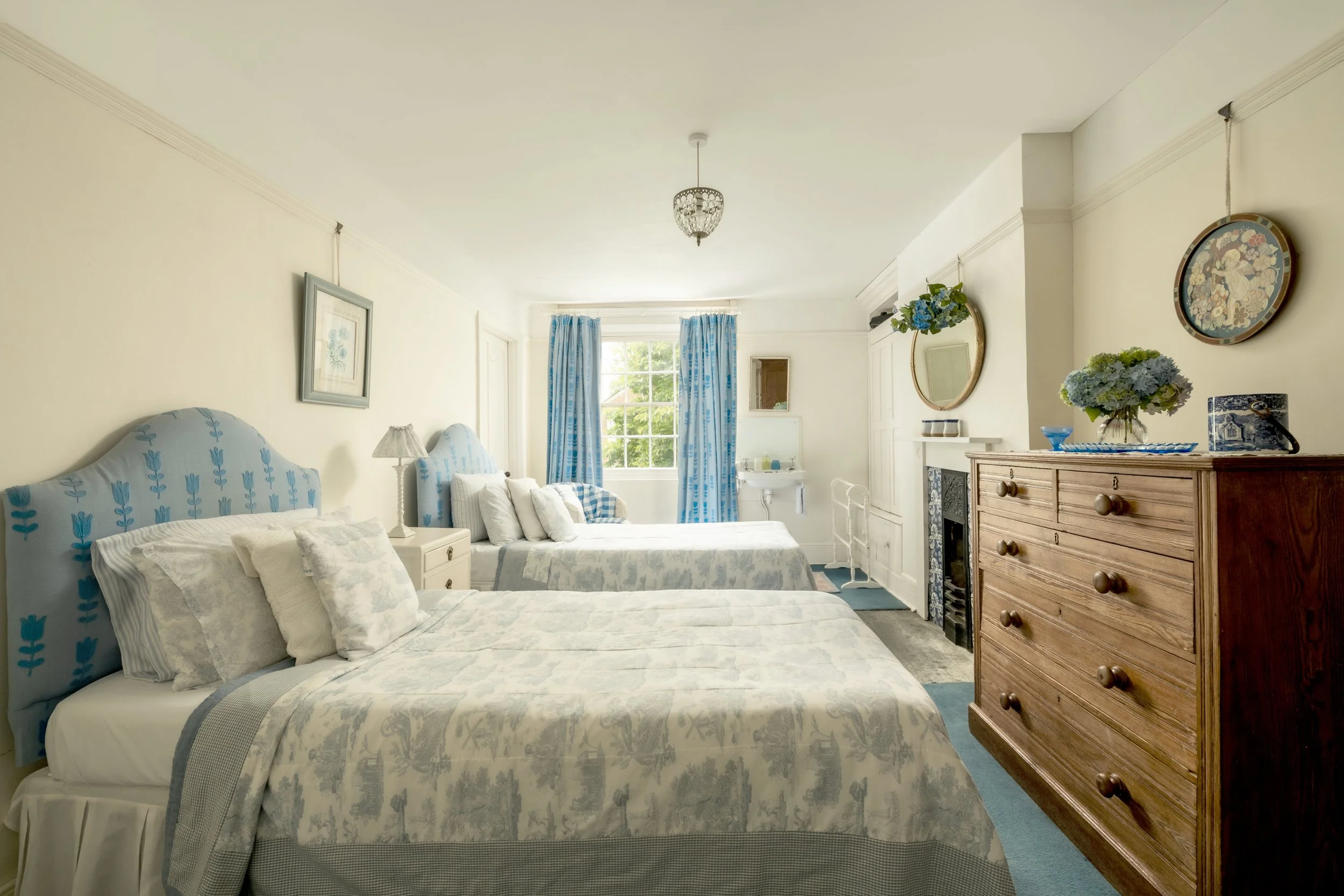 A cozy bedroom with three beds, each with white bedding and multiple pillows. The beds have blue and white patterned headboards. A wooden dresser with decorative items and a bouquet of flowers is on the right. A window with blue and white curtains al