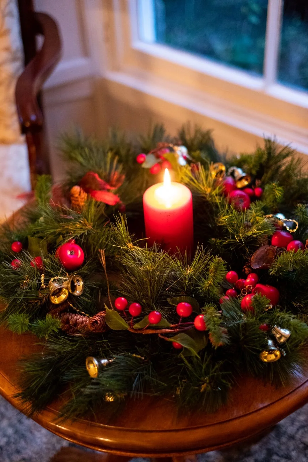 A decorated Christmas wreath with a lit red candle in the center, adorned with red berries, pine cones, and small gold and silver bells, on a wooden table near a window at Parnacott house.