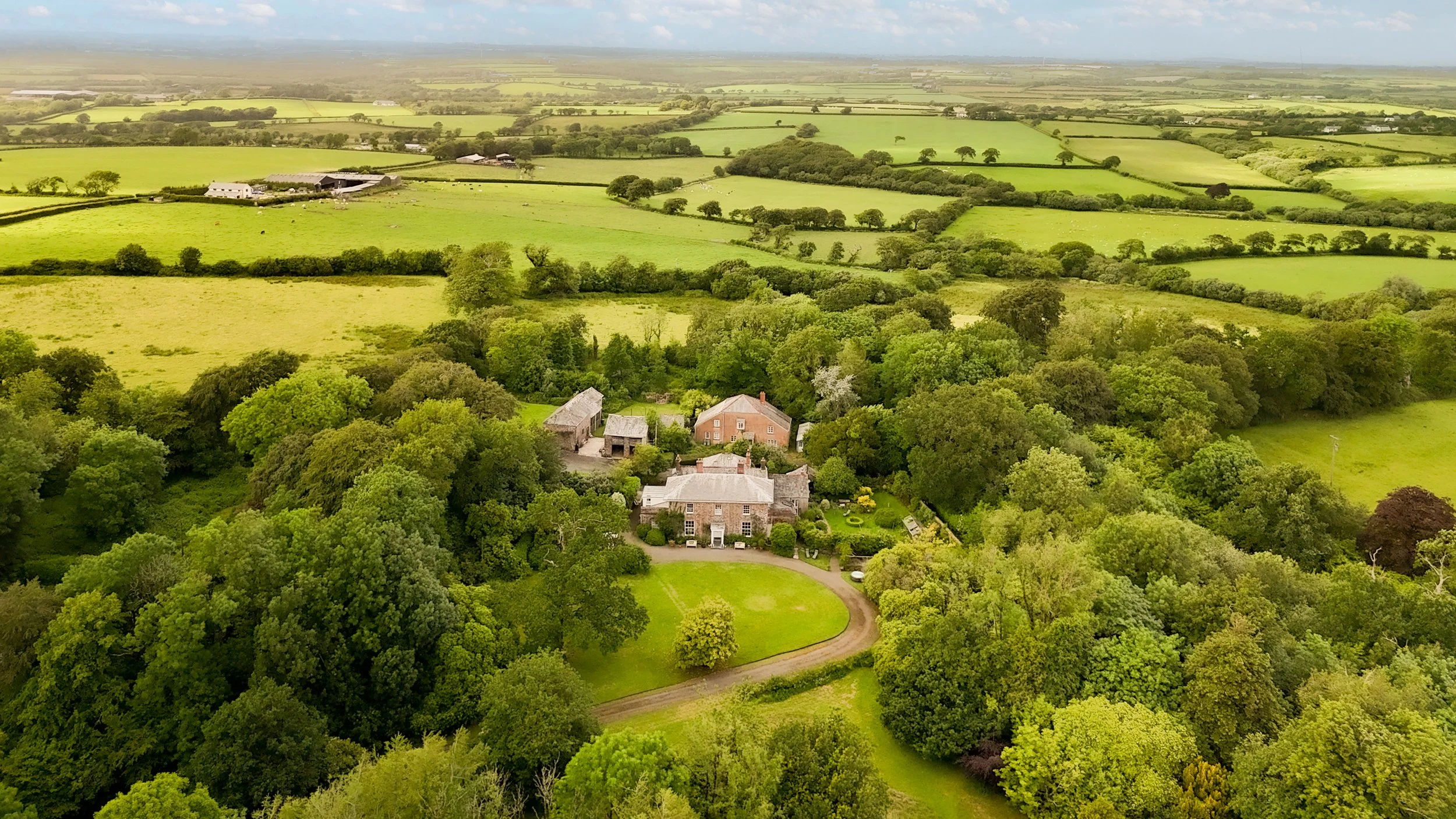 Aerial view of lush green farmland with scattered houses, barns, and a winding driveway surrounded by trees under a partly cloudy sky at Parnacott house.