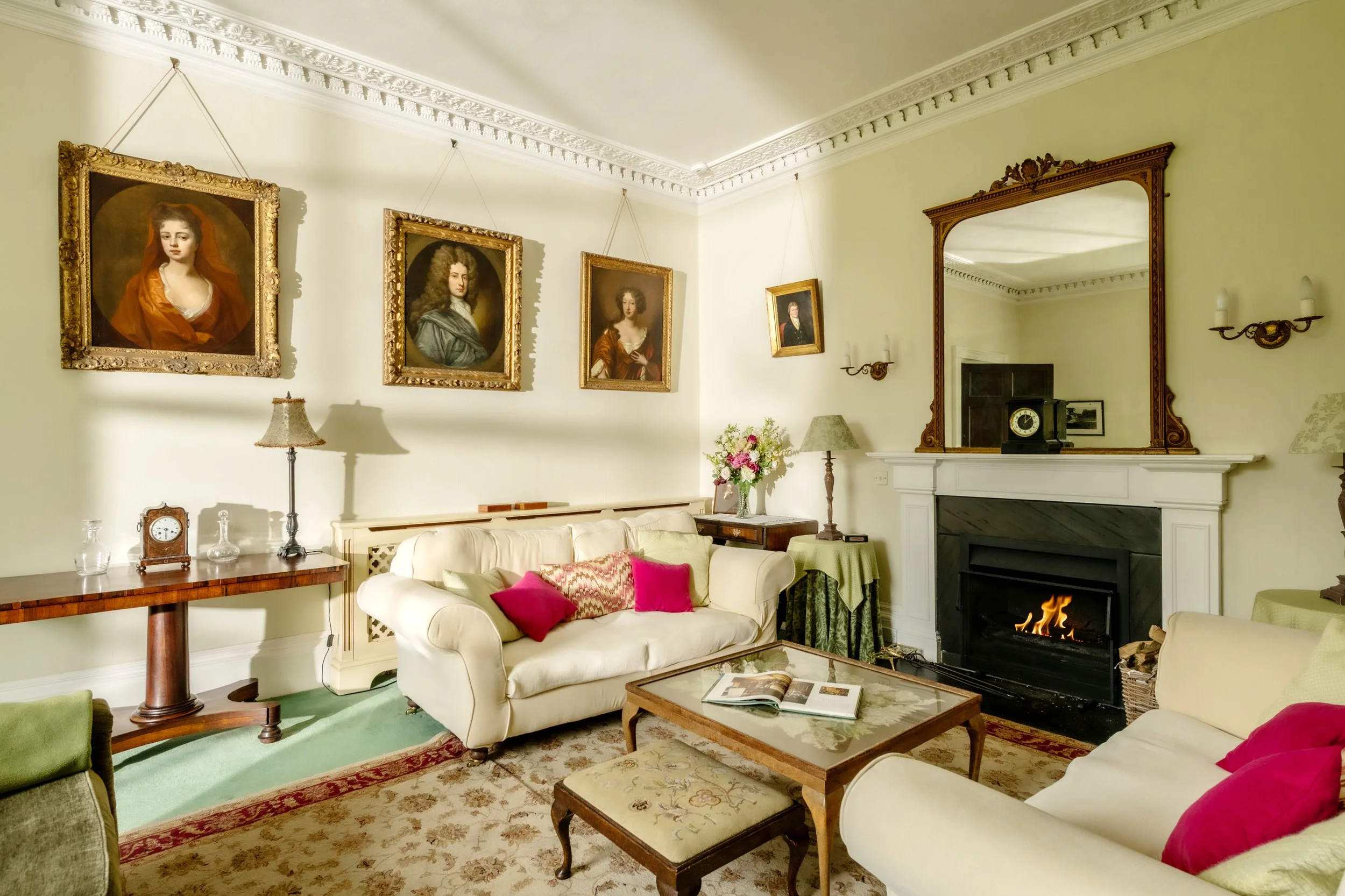 Living room with cream sofas, pink and green pillows, wooden coffee table, fireplace with mirror above, paintings on the wall, and decorative lighting fixtures at Parnacott house.