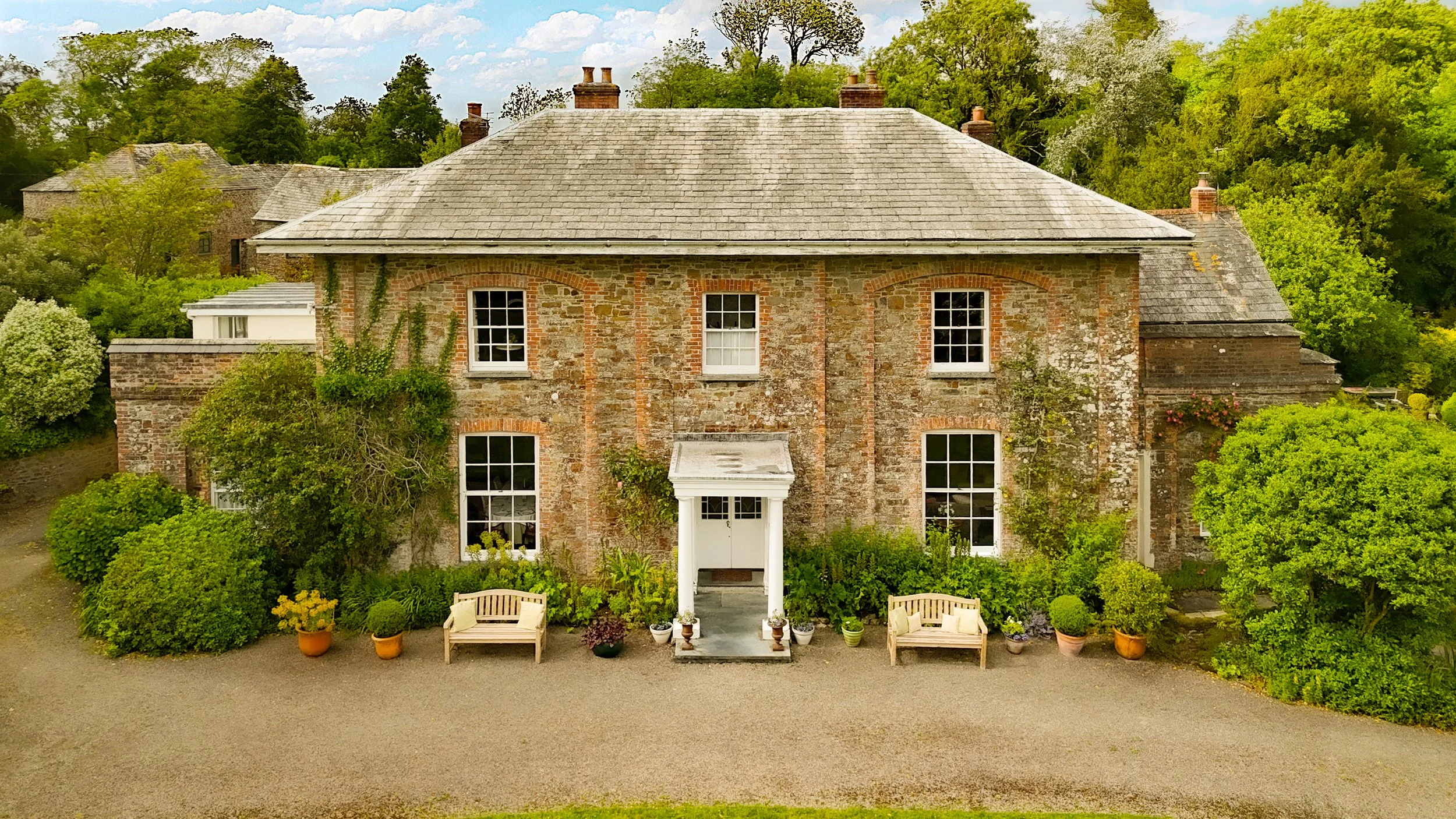 A large brick two-story house with a pitched roof, surrounded by greenery & potted plants in front. The house has multiple white-framed windows and an entryway with a small canopy. There are trees & other houses in the background at Parnacott house.