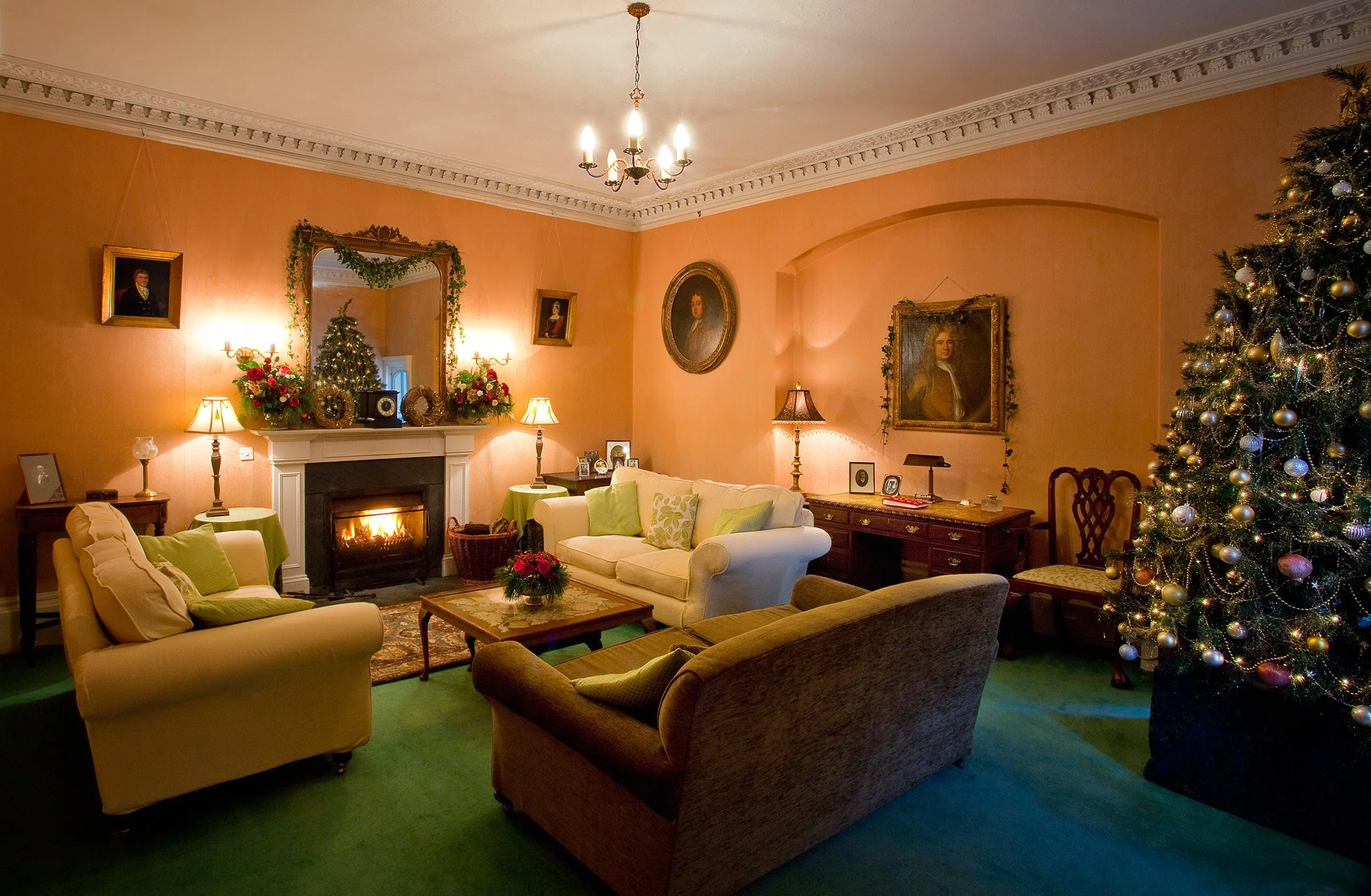 Living room decorated for Christmas with a large Christmas tree adorned with ornaments and lights, a fireplace with a mirror above it, several framed portraits on the walls, lamps, a white sofa with green pillows, a coffee table with holiday decorations, and classic furniture.