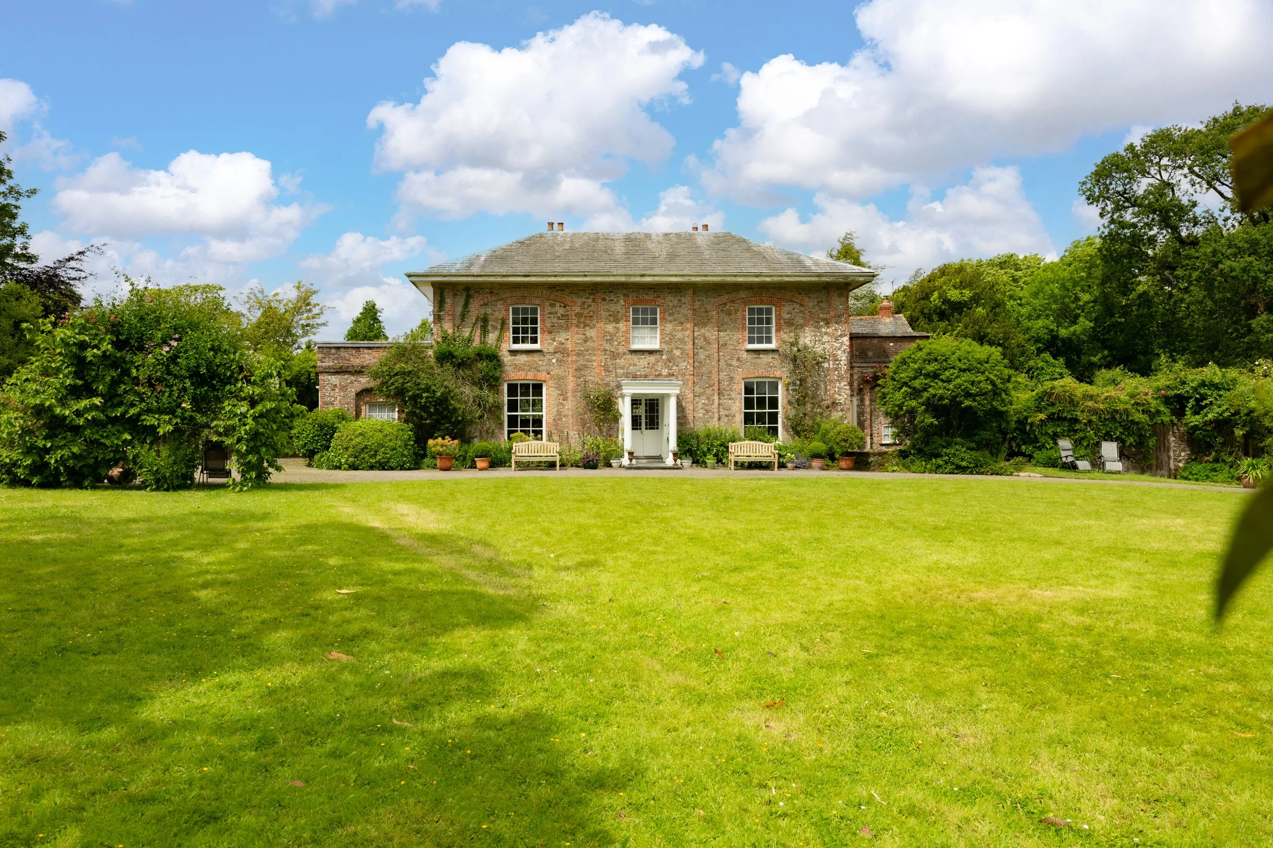 A two-story brick house with a gray roof, surrounded by a well-kept lawn and lush green trees and bushes, under a bright blue sky with scattered white clouds at Parnacott house.
