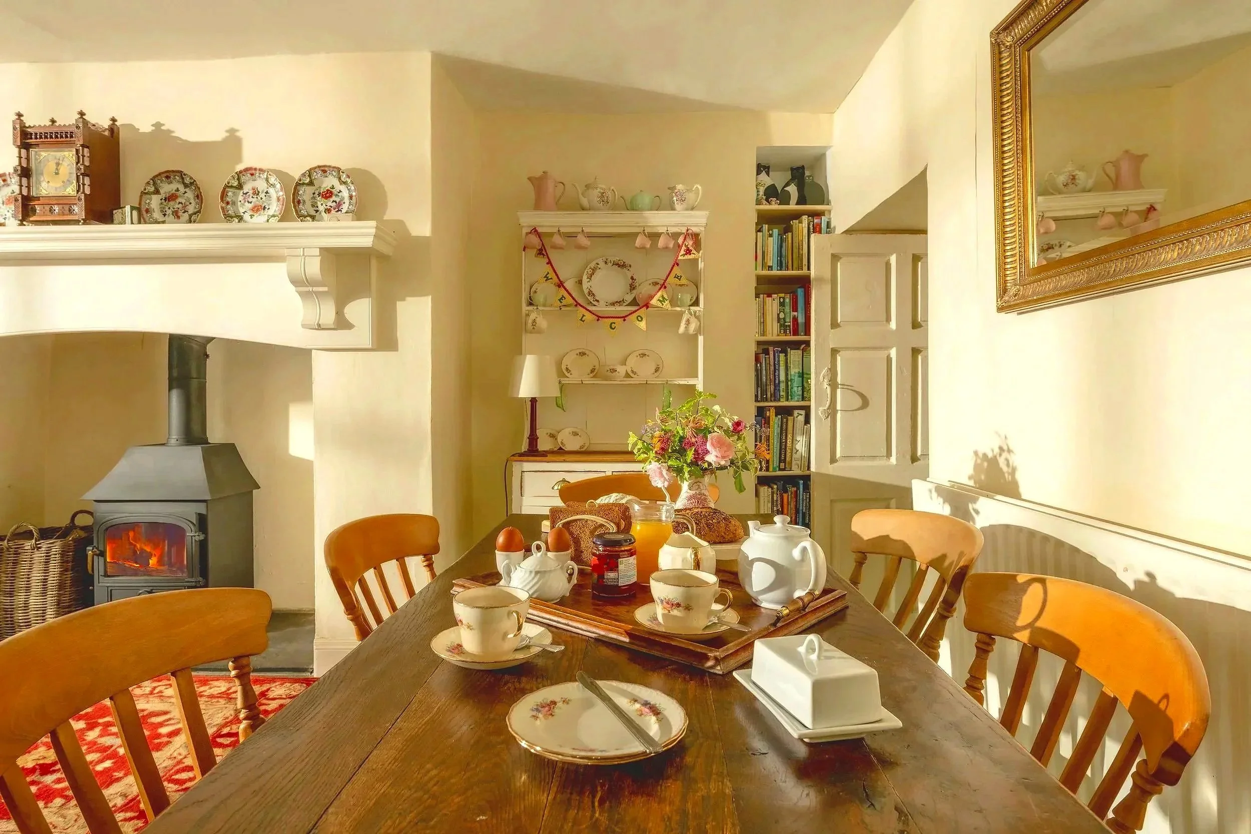 A cozy kitchen and dining area with a wood table set for breakfast, featuring cups, a teapot, and breakfast foods. The background includes shelves with decorative plates and books, a framed mirror, and a wood-burning stove at Parnacott house.
