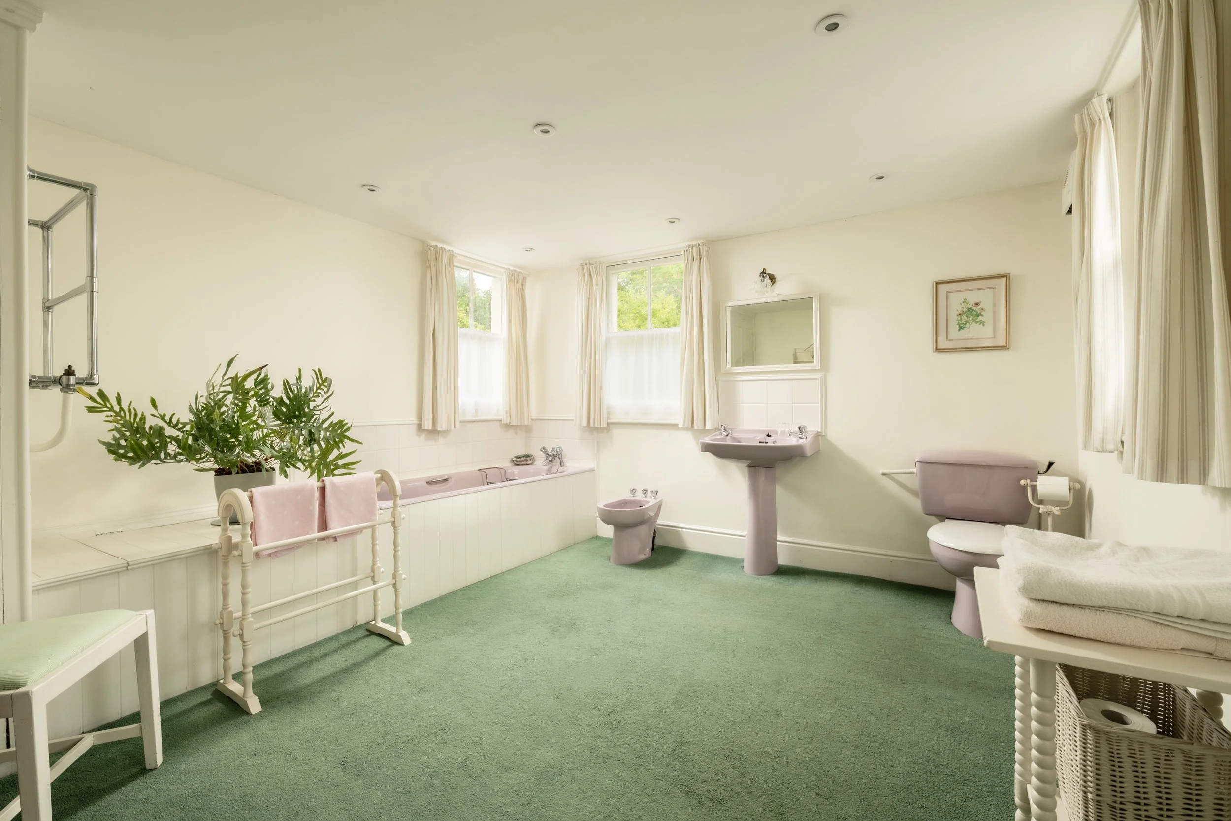 A spacious bathroom with cream-colored walls and a green carpet. It features a bathtub under three large windows with white curtains, a sink with a mirror, a toilet, and a bidet. There is a framed floral picture on the wall, a potted plant, and some 