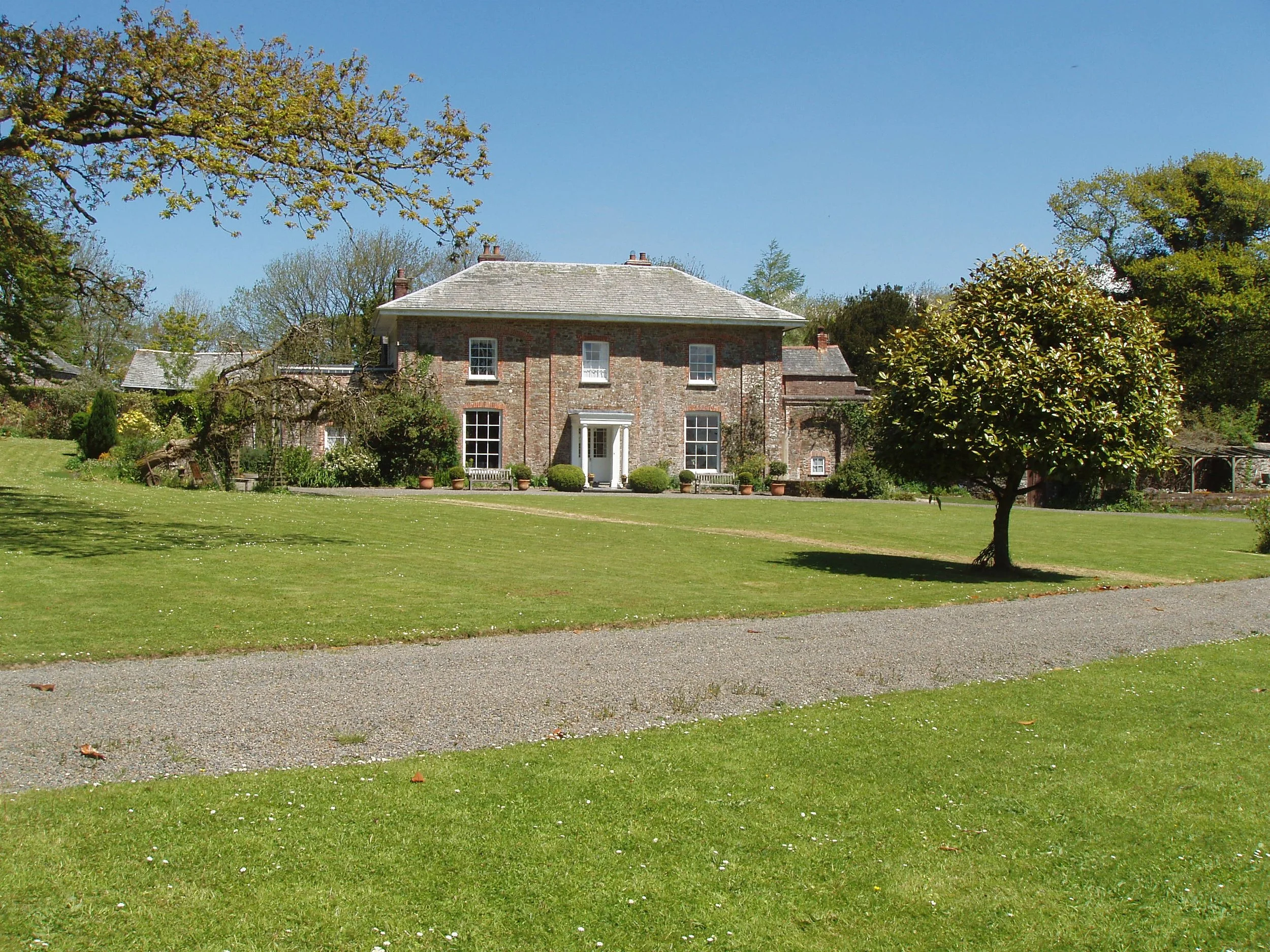 A large brick house with a grey shingle roof, surrounded by a well-maintained lawn, trees, and potted plants, under a clear blue sky Parnacott house. Parnacott Paws.