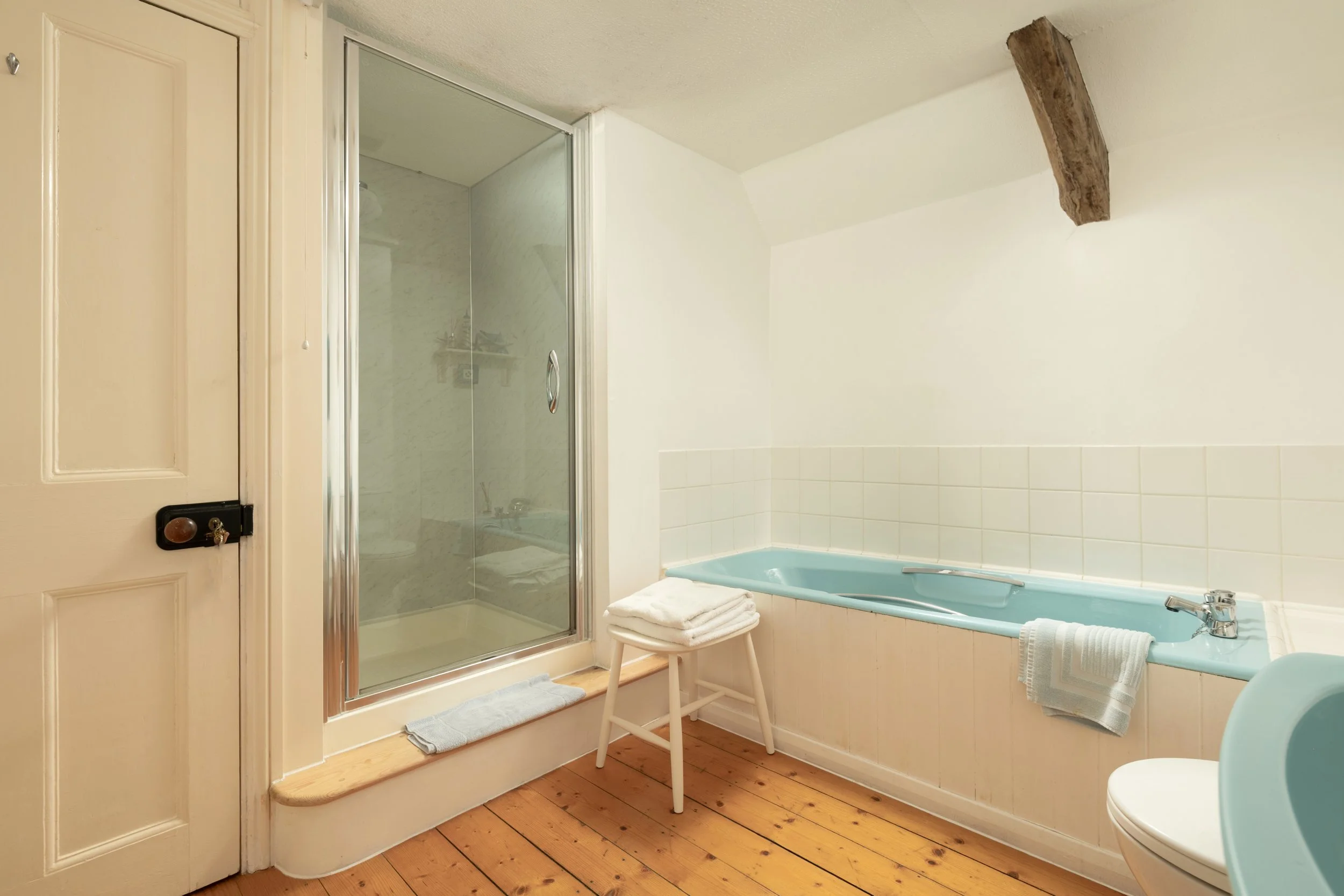 A bathroom with a shower, bathtub, and wooden floor. Towels are placed near the bathtub, and a stool with towels is next to the shower. There is a wooden beam on the ceiling and a closed door to the left at Parnacott house. 