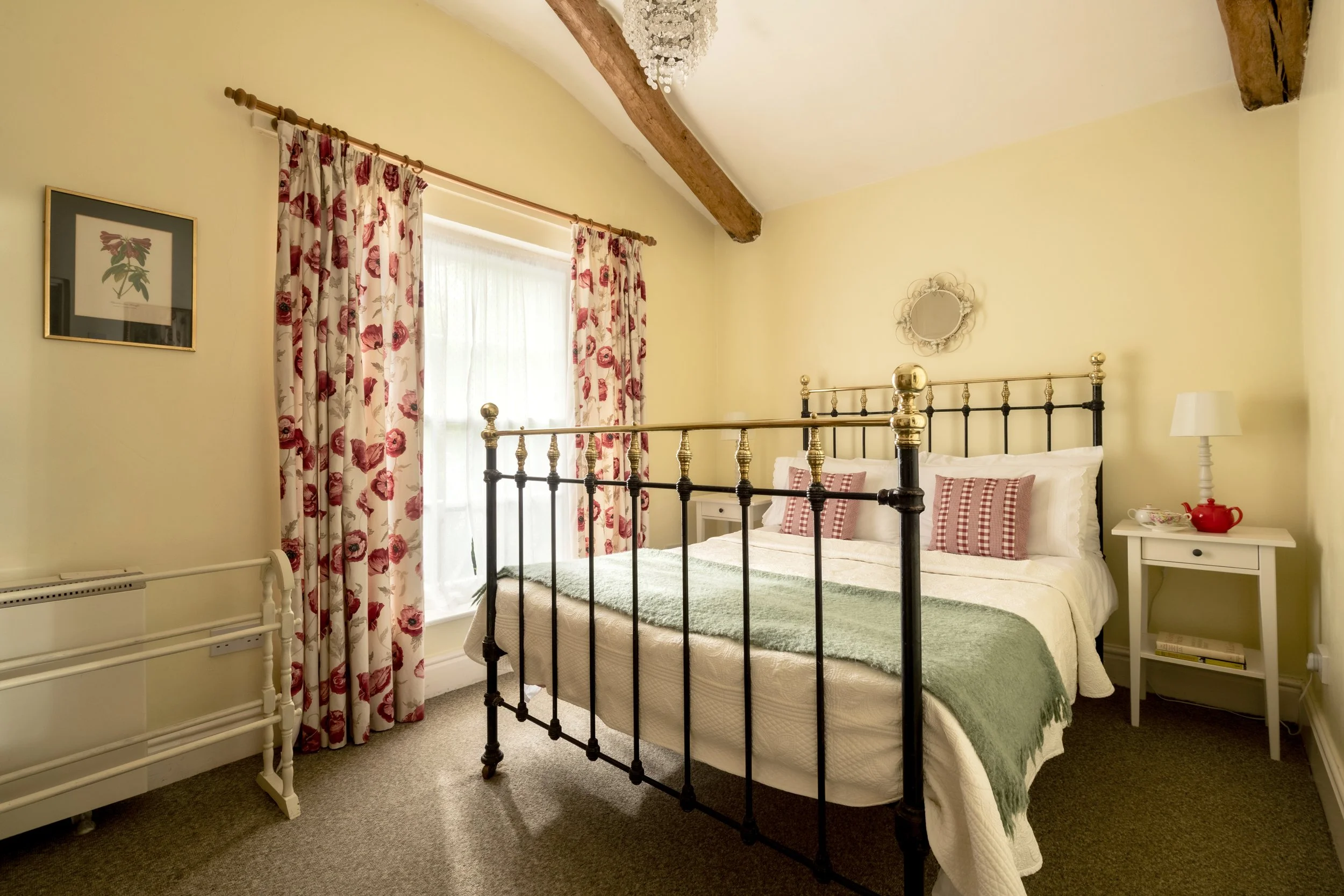 A cozy bedroom with a black metal bed frame, white bedding, and two red and white checked pillows. A bedside table with a white lamp and a red teapot is on the right. The window has floral curtains with red flowers and cream background, and is decora