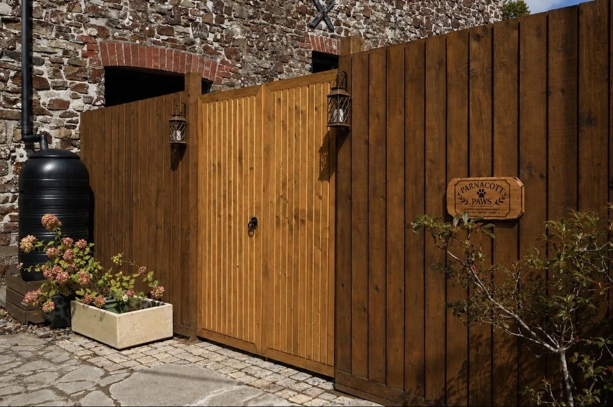 Wooden fence with a gate, a sign reading "Parnacott Paws," and potted plants with pink flowers. Parnacott house. Parnacott Paws.