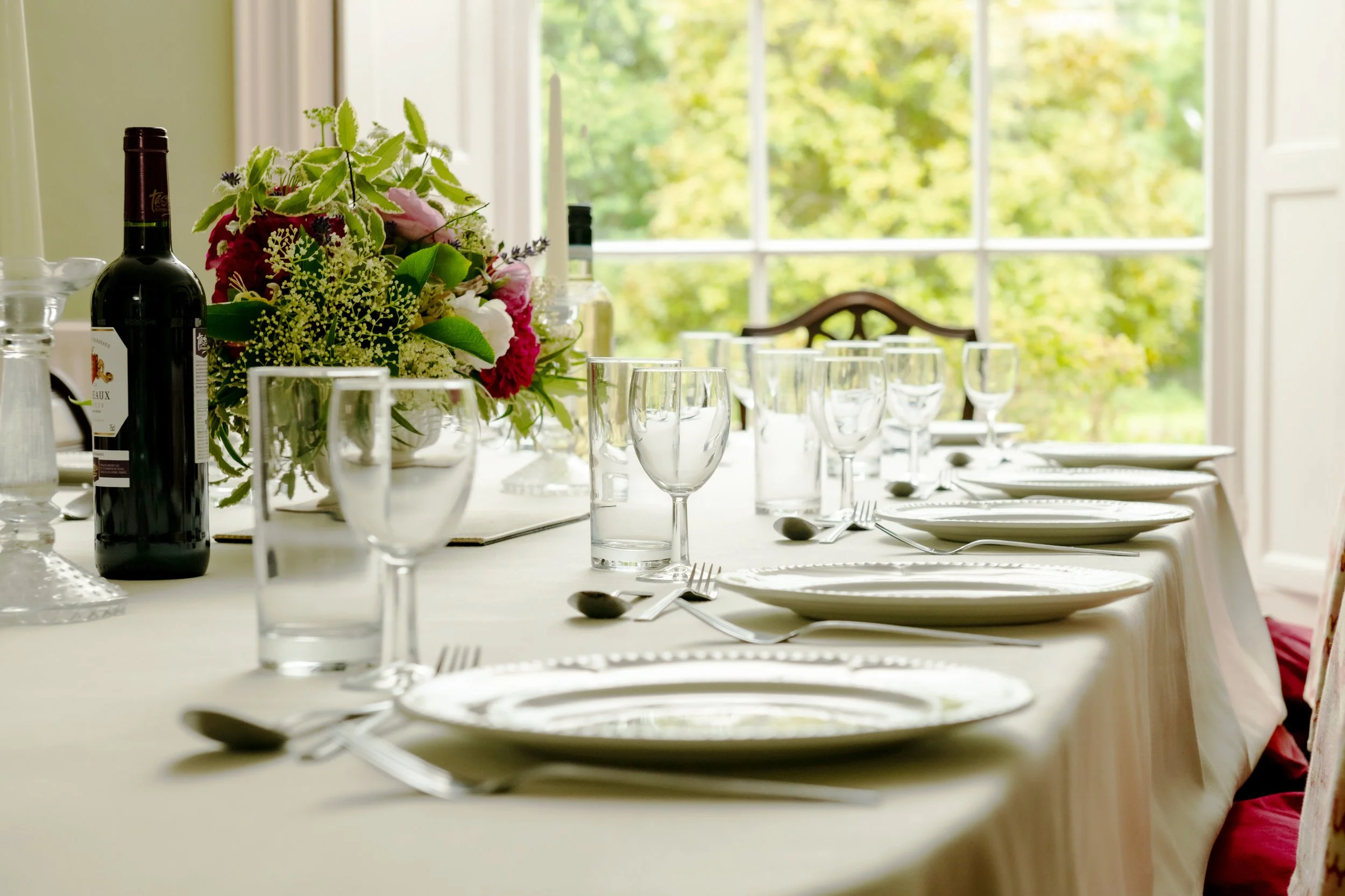 A banquet table set with white plates, silverware, wine glasses, and a floral centerpiece in front of a large window with a view of green trees at Parnacott house.