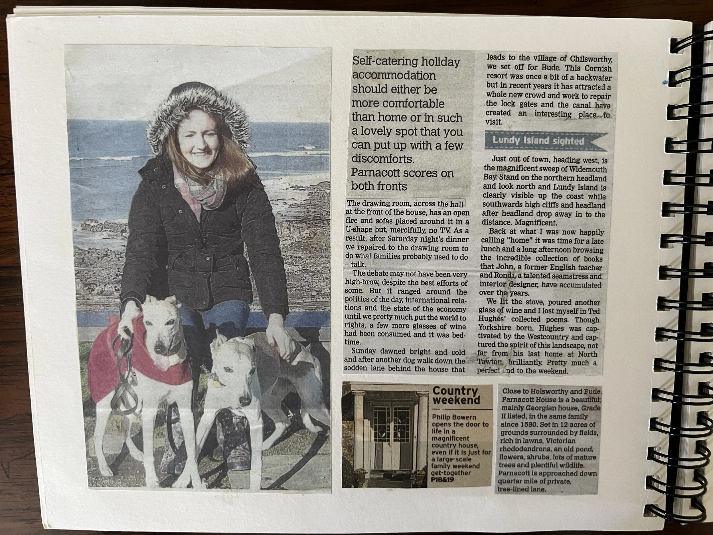 A woman with long hair wearing a winter coat and a fur-lined hat, smiling, with two dogs outdoors near a rocky shoreline and ocean in the background at Bude, near Parnacott house.