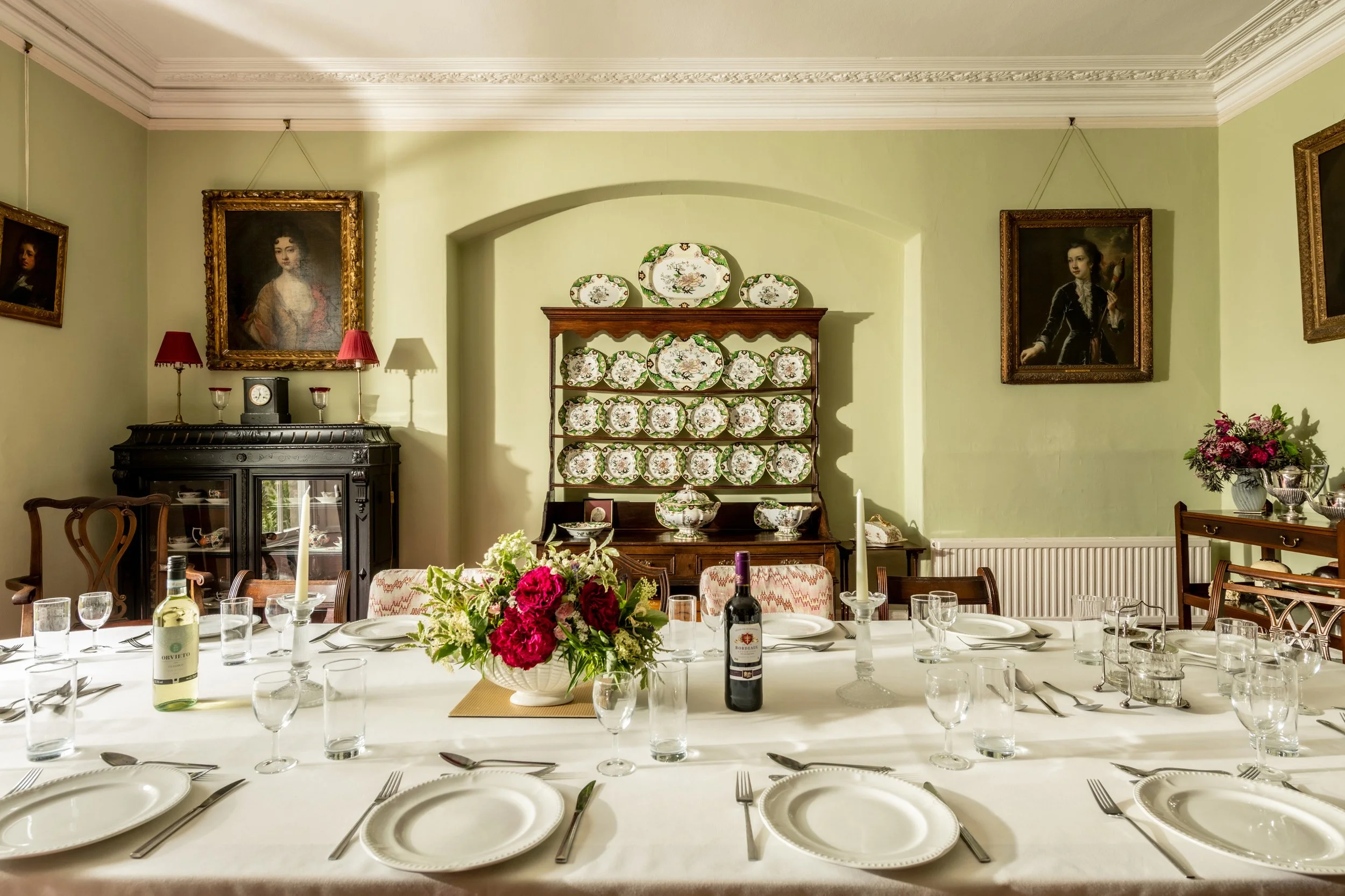 A dining room set up for a formal meal with a long white tablecloth, glasses, plates, and cutlery, and a centerpiece of pink and purple flowers. The room has pale green walls, ornate crown molding, and framed portraits hanging on the walls. A china cabinet with decorative plates is in the background