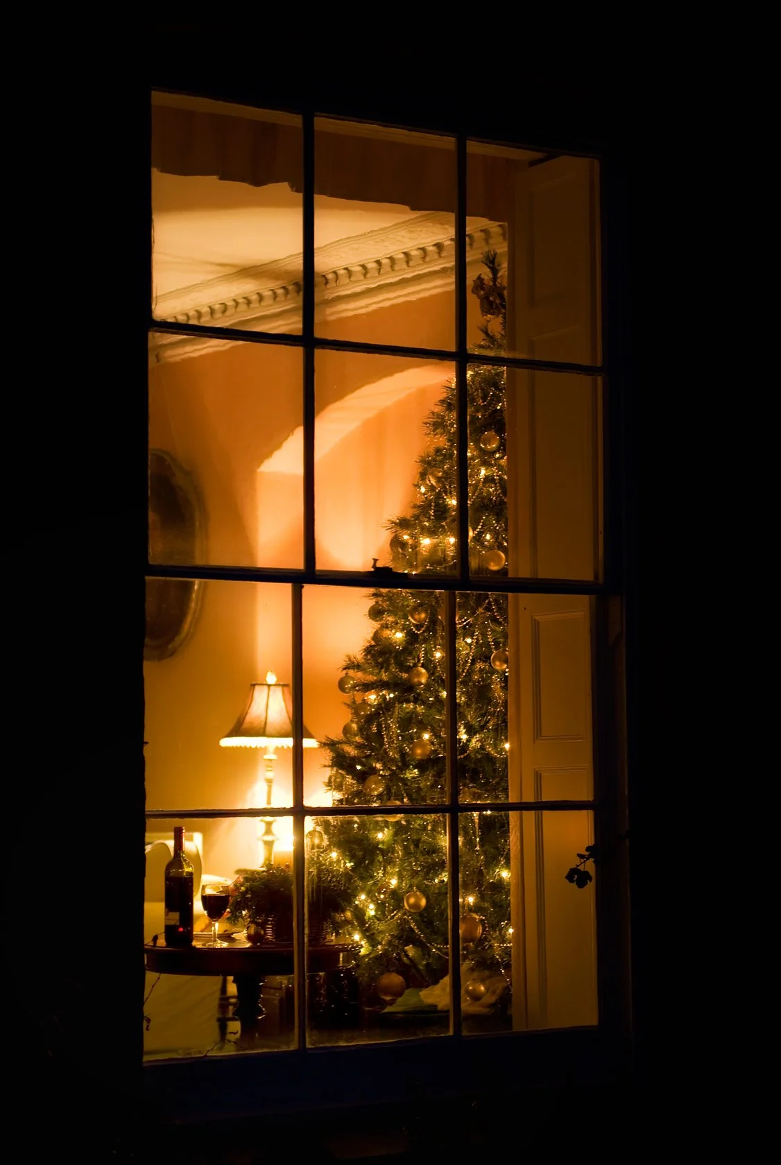 View through a window showing a decorated Christmas tree with lights and ornaments inside a cozy room, with a lamp and a table holding a bottle of wine and a glass at Parnacott house.